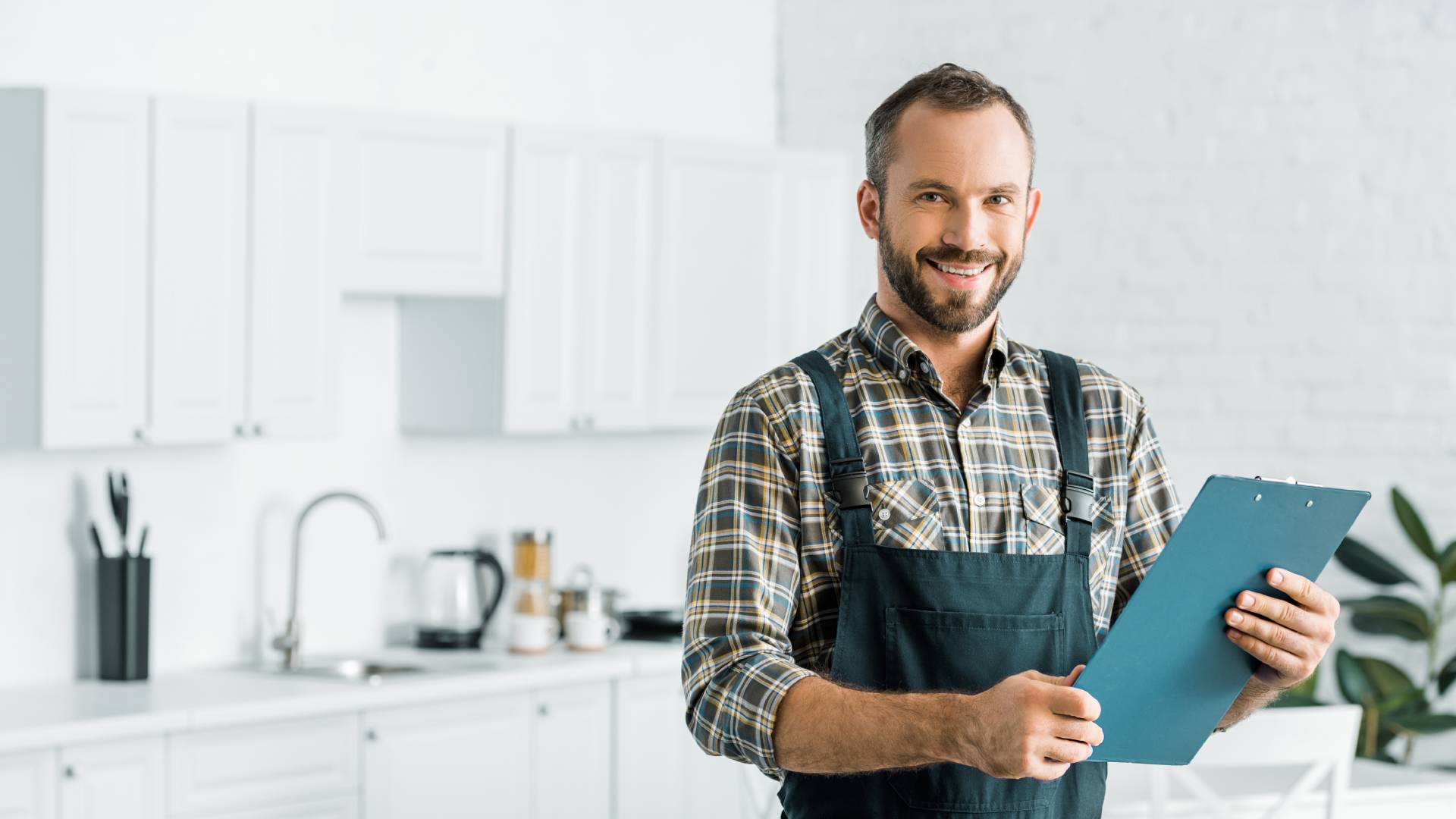A smiling plumber wearing a tool belt, standing in a bright residential kitchen, and holding a blue clipboard with both hands.