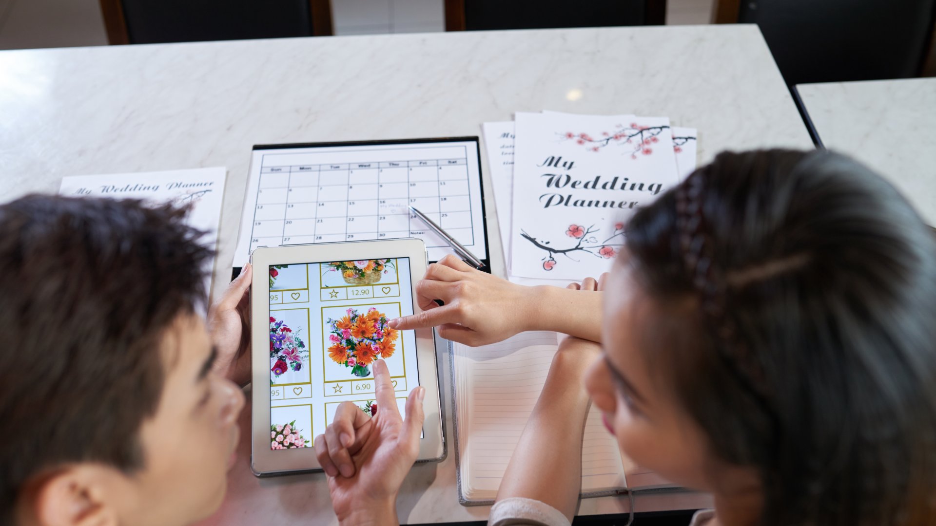 A man and woman pointing at a tablet with flower bouquet options beside an open wedding planner and calendar.