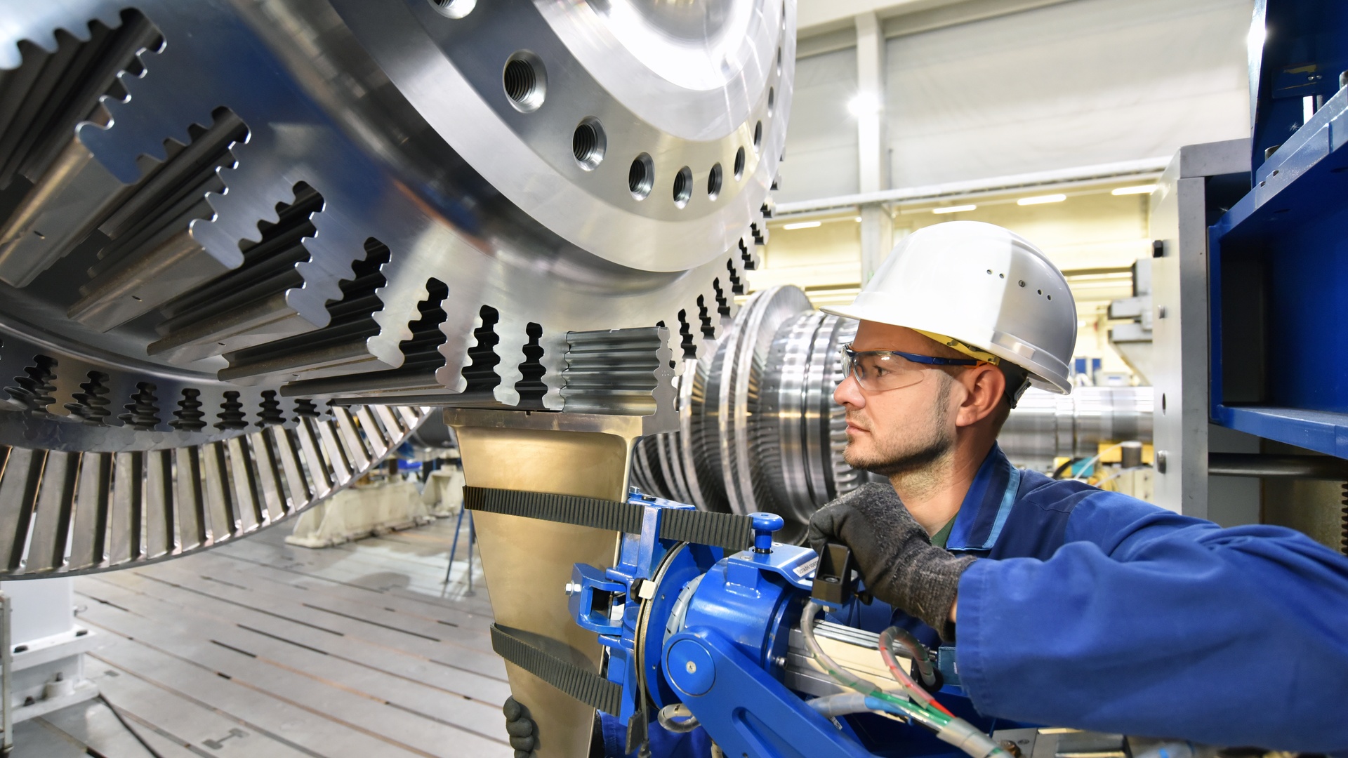 Close-up of an industrial worker working on a metal gas turbine in a factory. The worker has safety equipment on.
