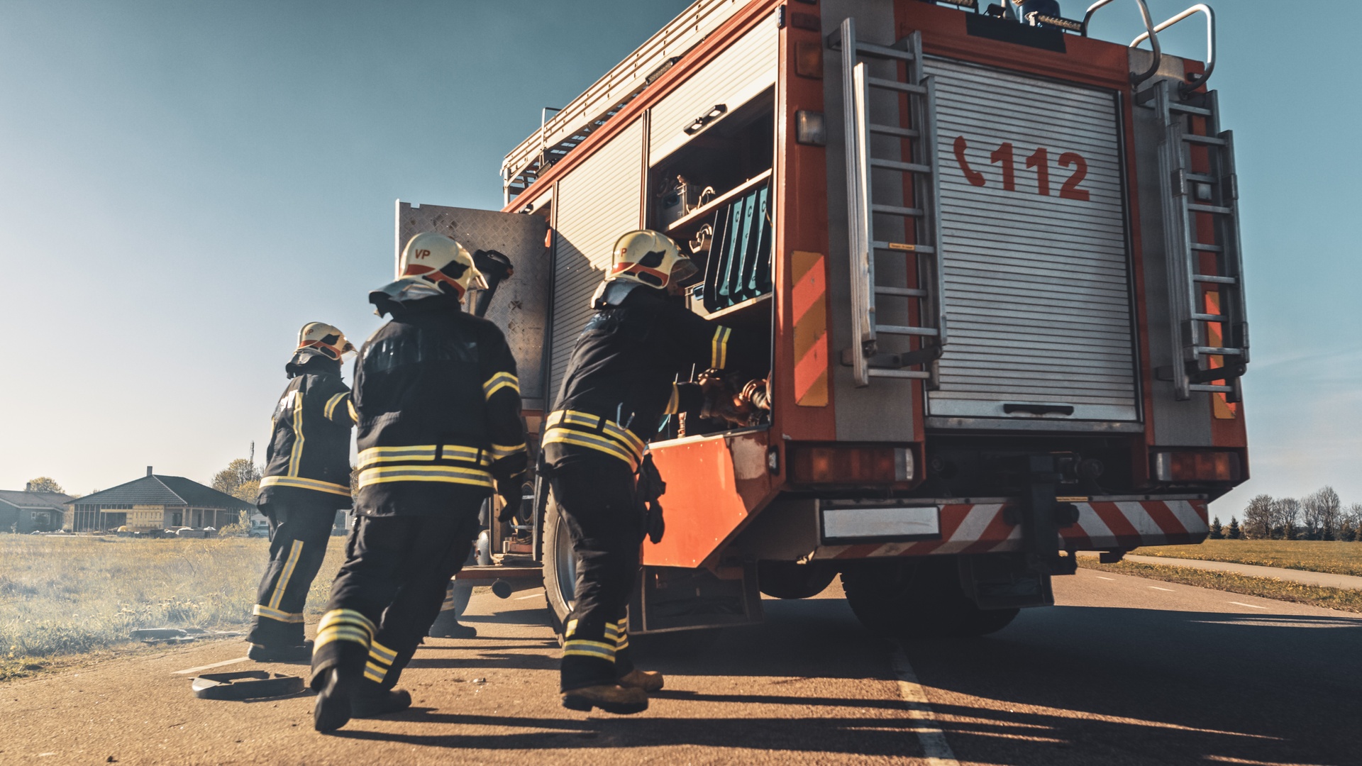 Three emergency responders dressed in uniform are grabbing tools from a red fire truck that sits on a road.