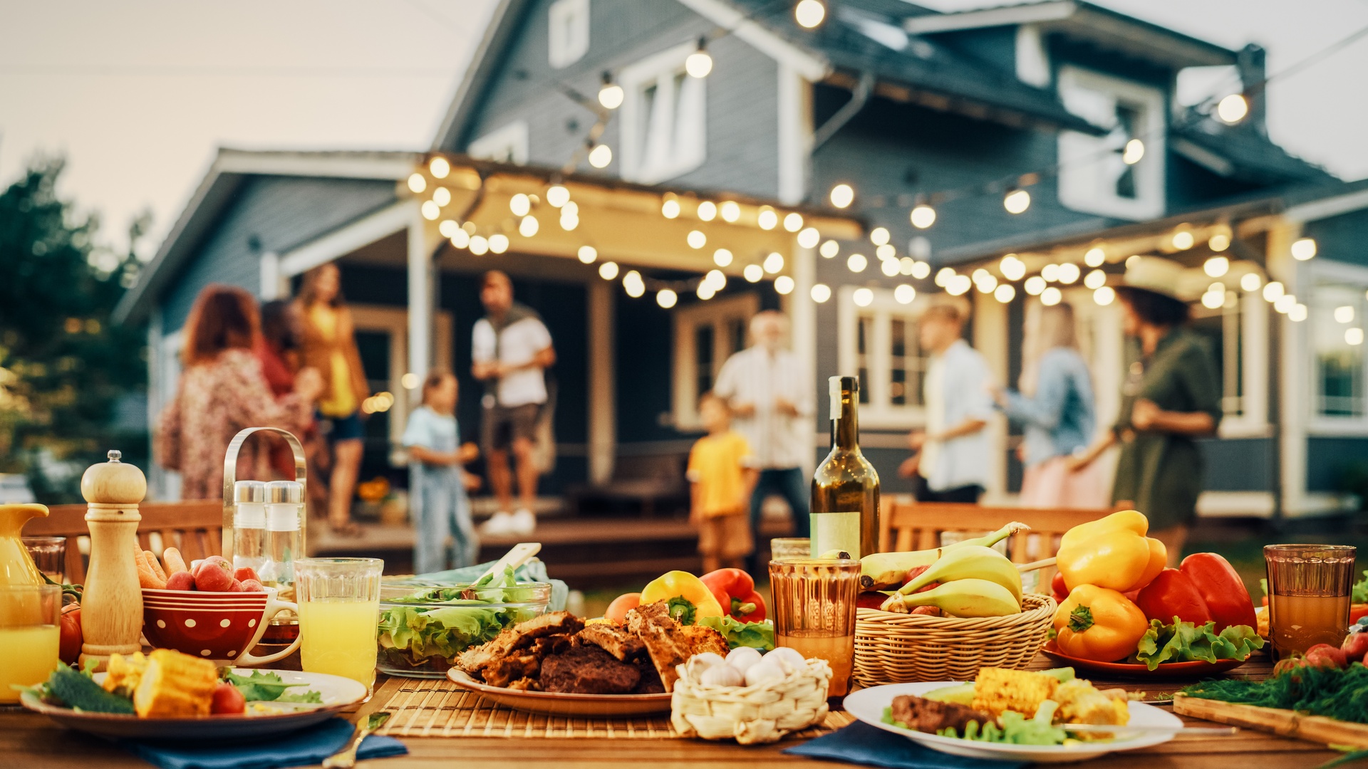 A close-up of a dining table with multiple food and drink options placed in a backyard with people in the background.