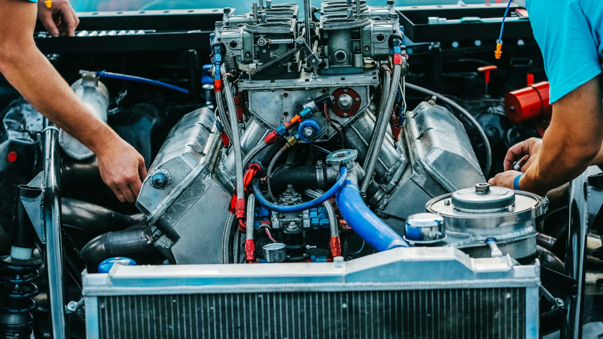 A close-up of the engine of a classic sports car with a massive engine, the hood open and two mechanics working on it.