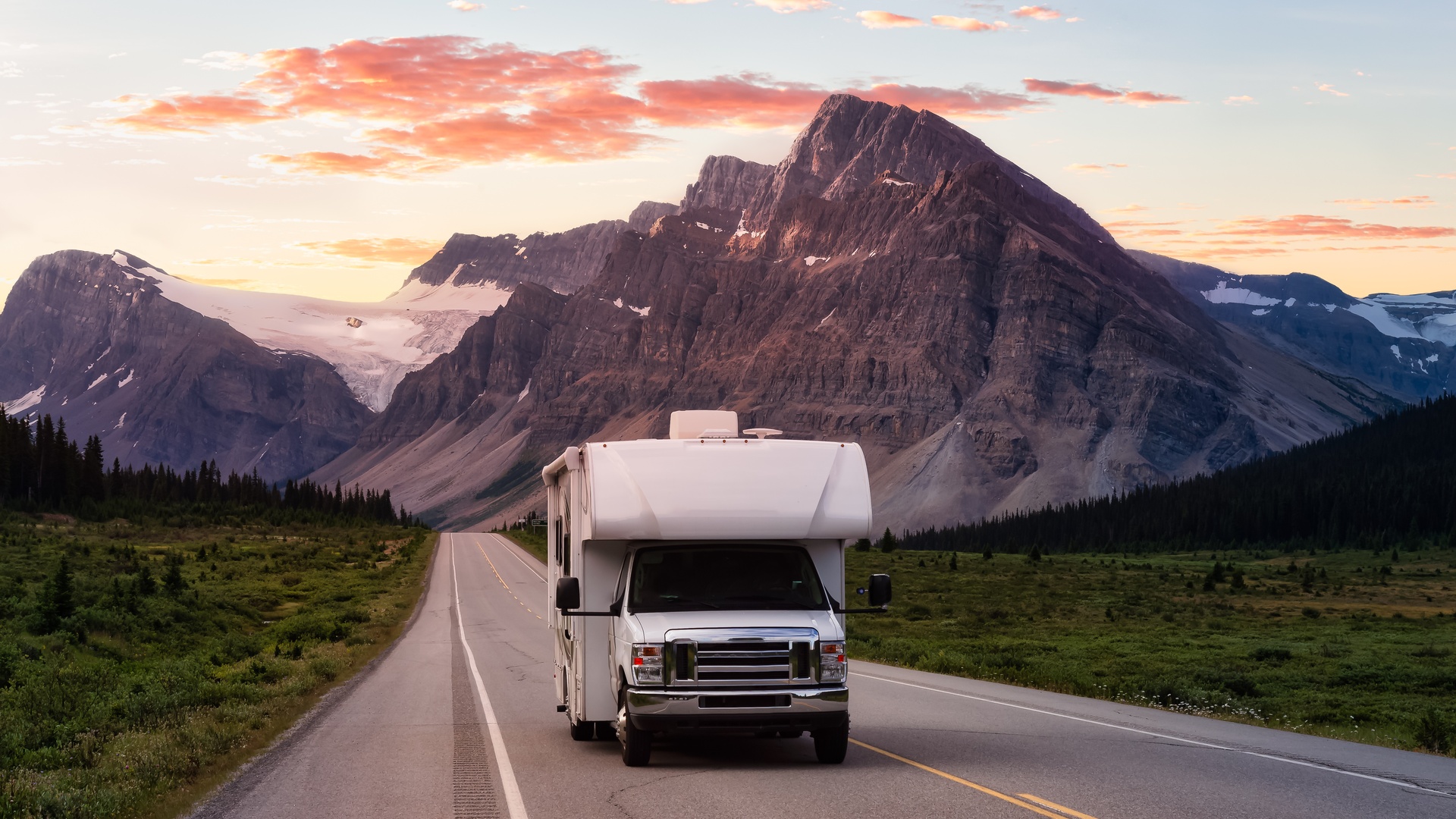 A white RV driving down an open road with a snowcapped mountain range and a sunset in the background.