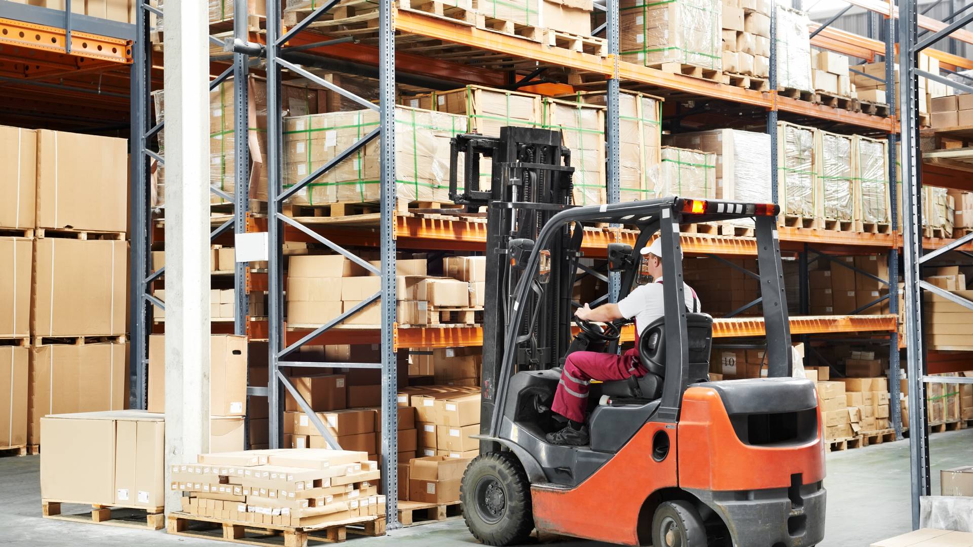 A warehouse employee wearing a white hard hat, maroon pants, and white shirt drives an orange and black forklift.