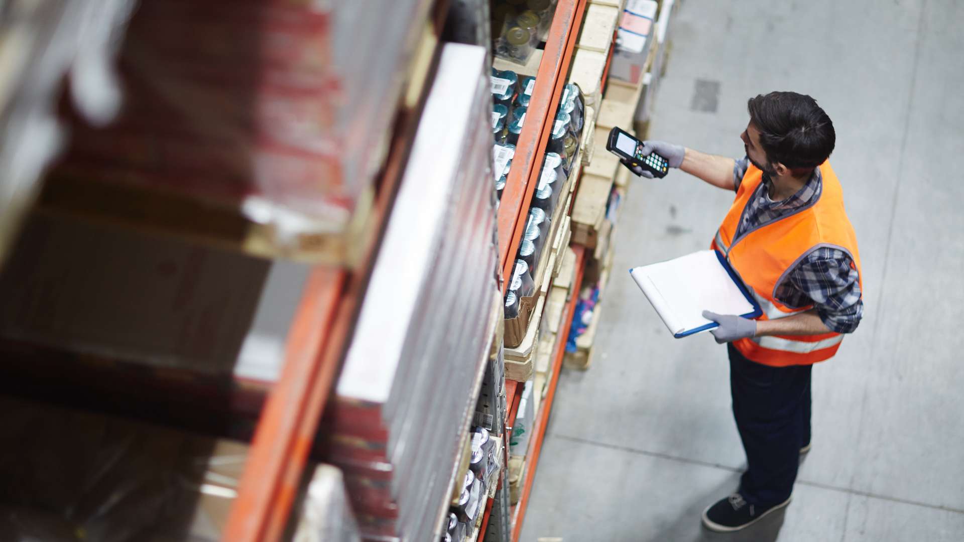 A warehouse worker is wearing an orange vest and holding a clipboard. They're holding a digital device up a box.