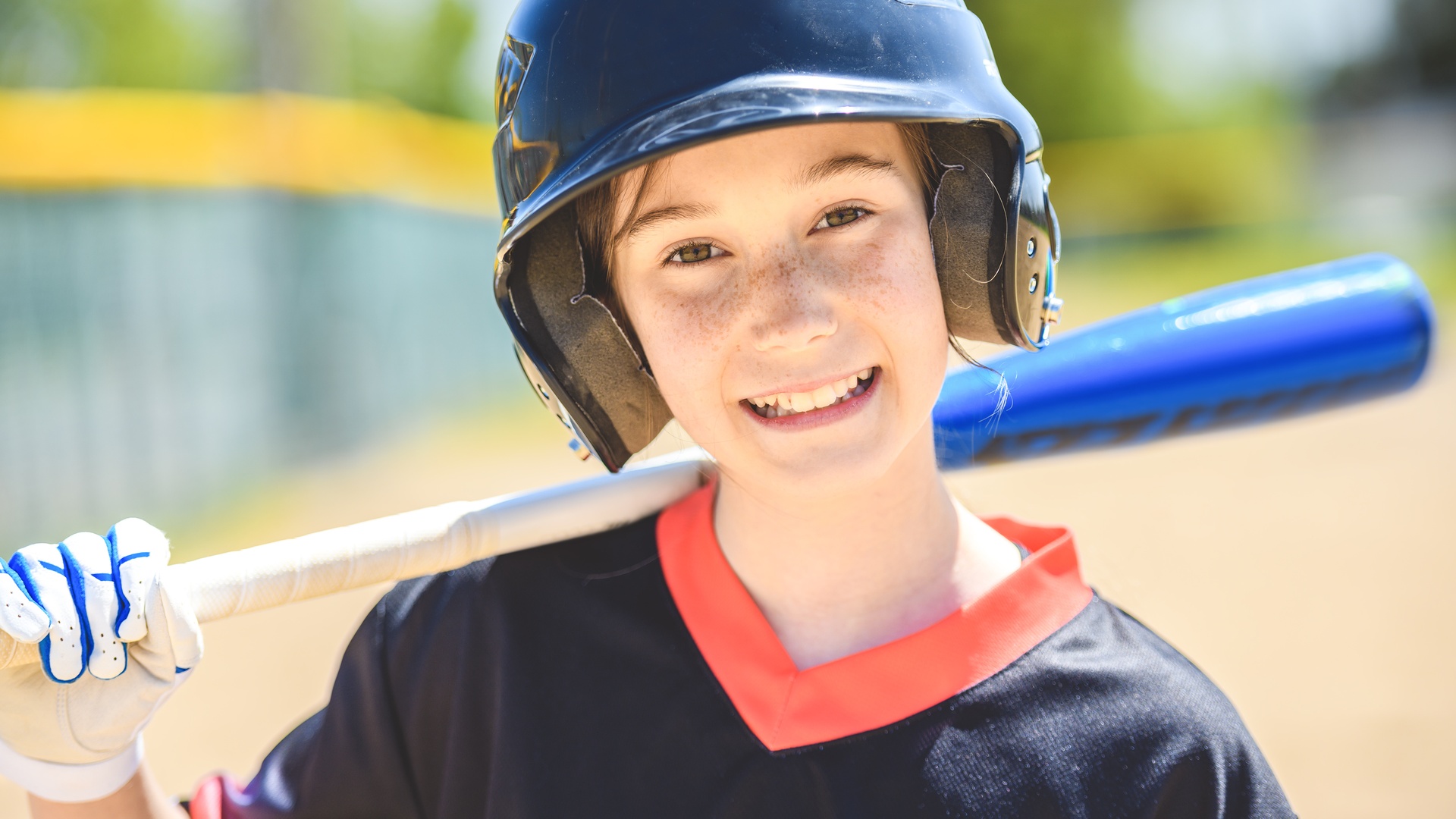A close-up view shows a child wearing a black safety helmet and a black shirt. The child holds a blue bat over their shoulder.