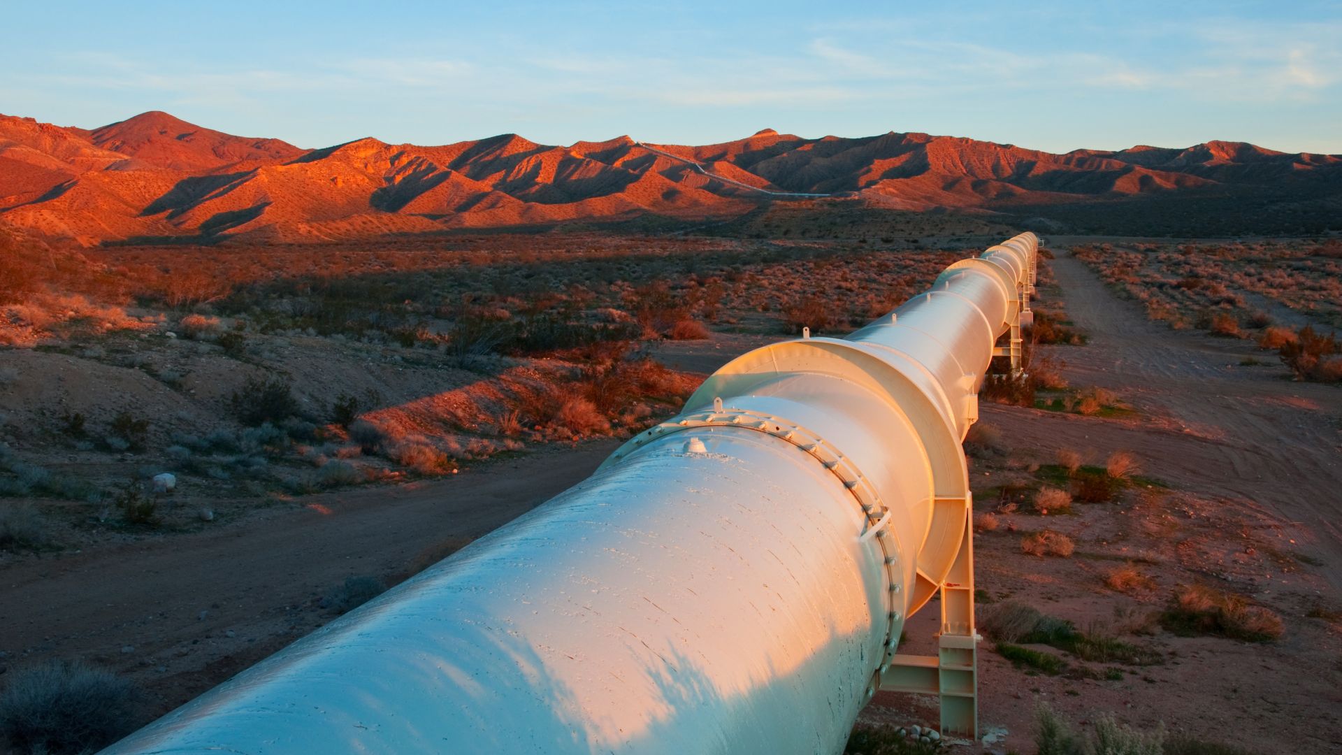 A large, white, elevated pipeline runs through a desert. Large mountains appear in the background.