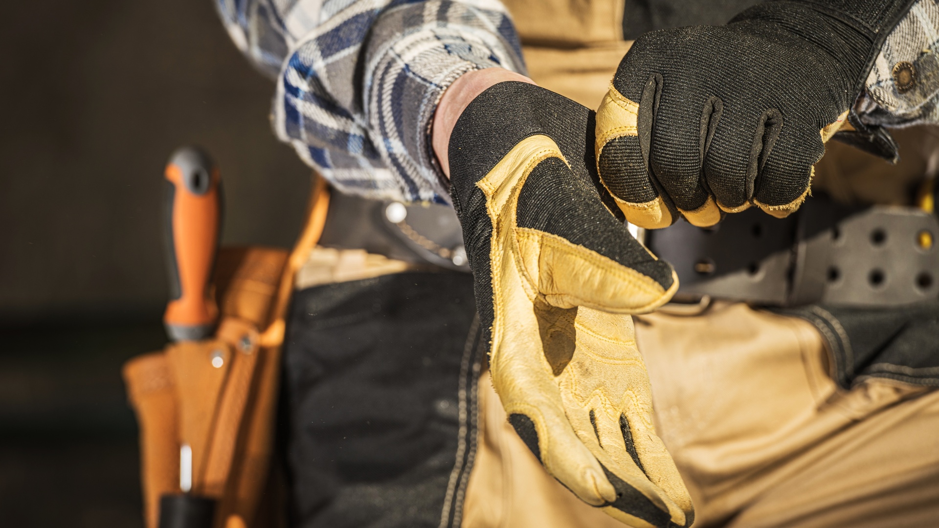 A close-up of a person wearing a plaid shirt and a tool belt putting on a pair of yellow work gloves.