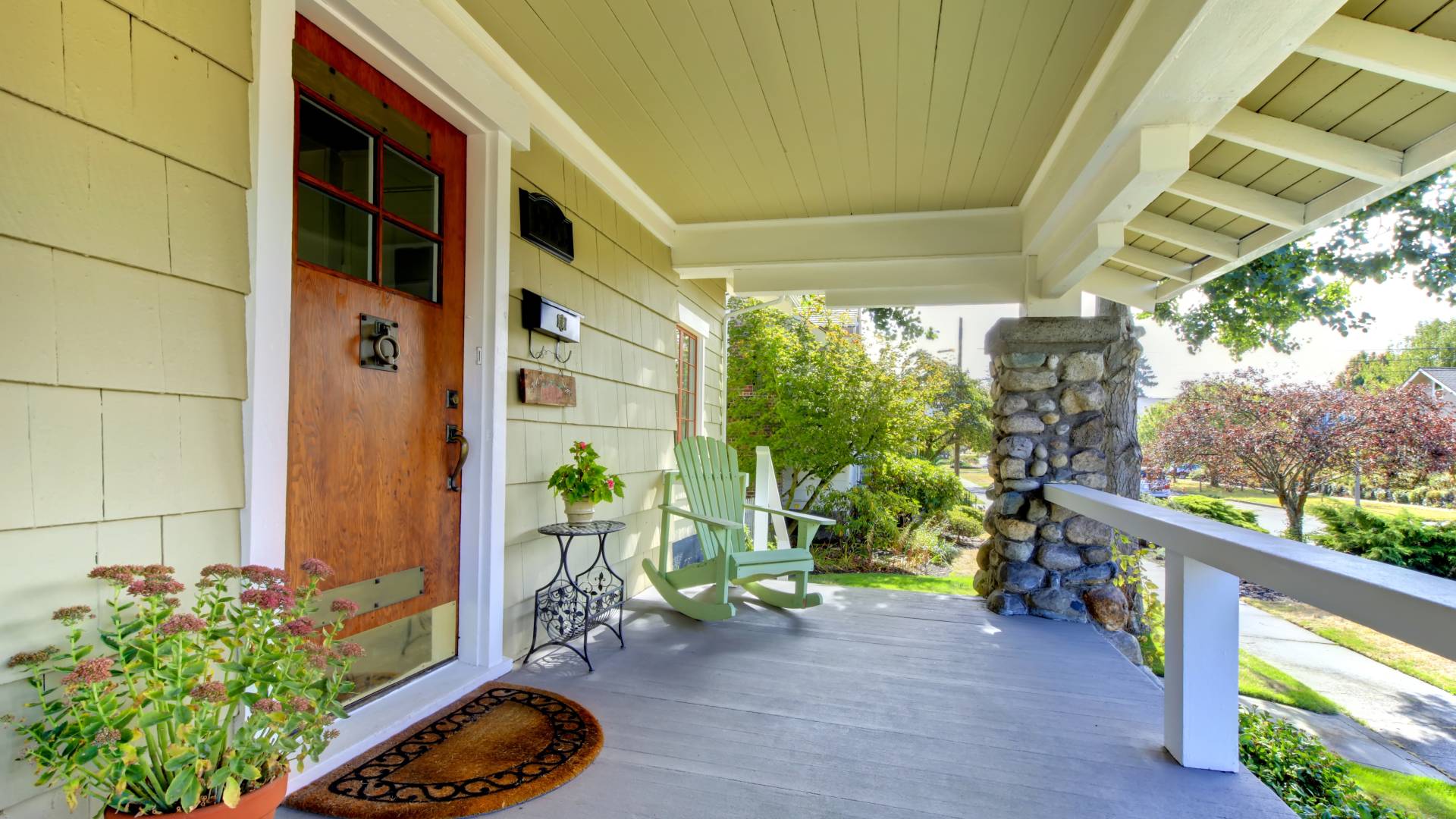 A home's front porch features a doormat, potted plants, and a rocking chair. The front door features brown paint.