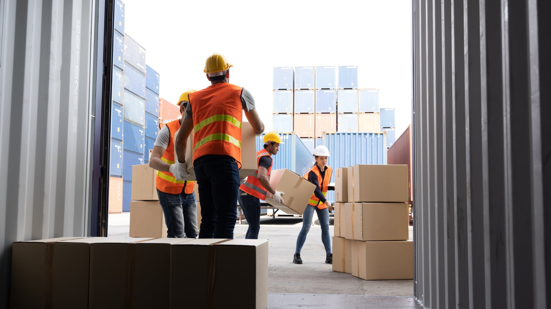 Four workers wearing bright orange vests load cardboard shipping cargo into a large shipping container.