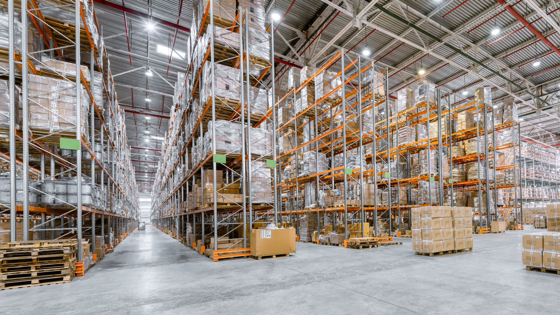 High bay lights are spaced evenly apart on a high warehouse ceiling. They evenly illuminate aisles and a large open space.