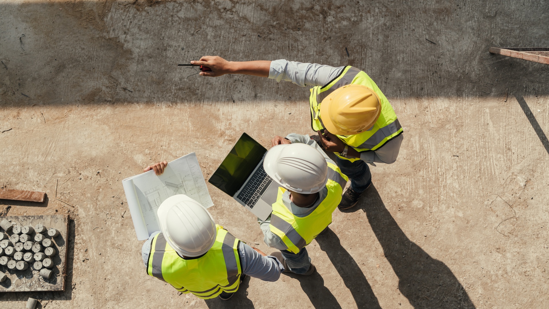 An overhead view of three men in protective construction gear. One points, one holds a laptop, and the other holds blueprints.