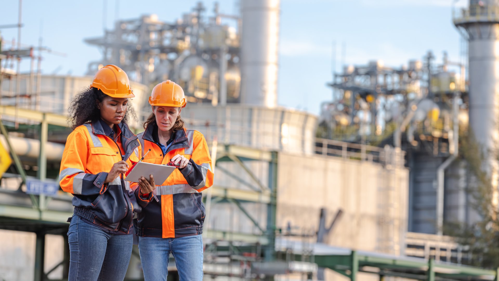 Two women wearing orange safety jackets and orange hard hats look at a tablet. Large pipelines are in the background.