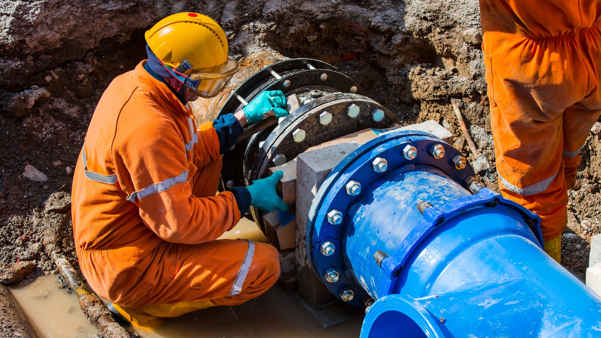 A man in an orange protective jumpsuit squats next to an exposed blue industrial pipeline to perform repairs.
