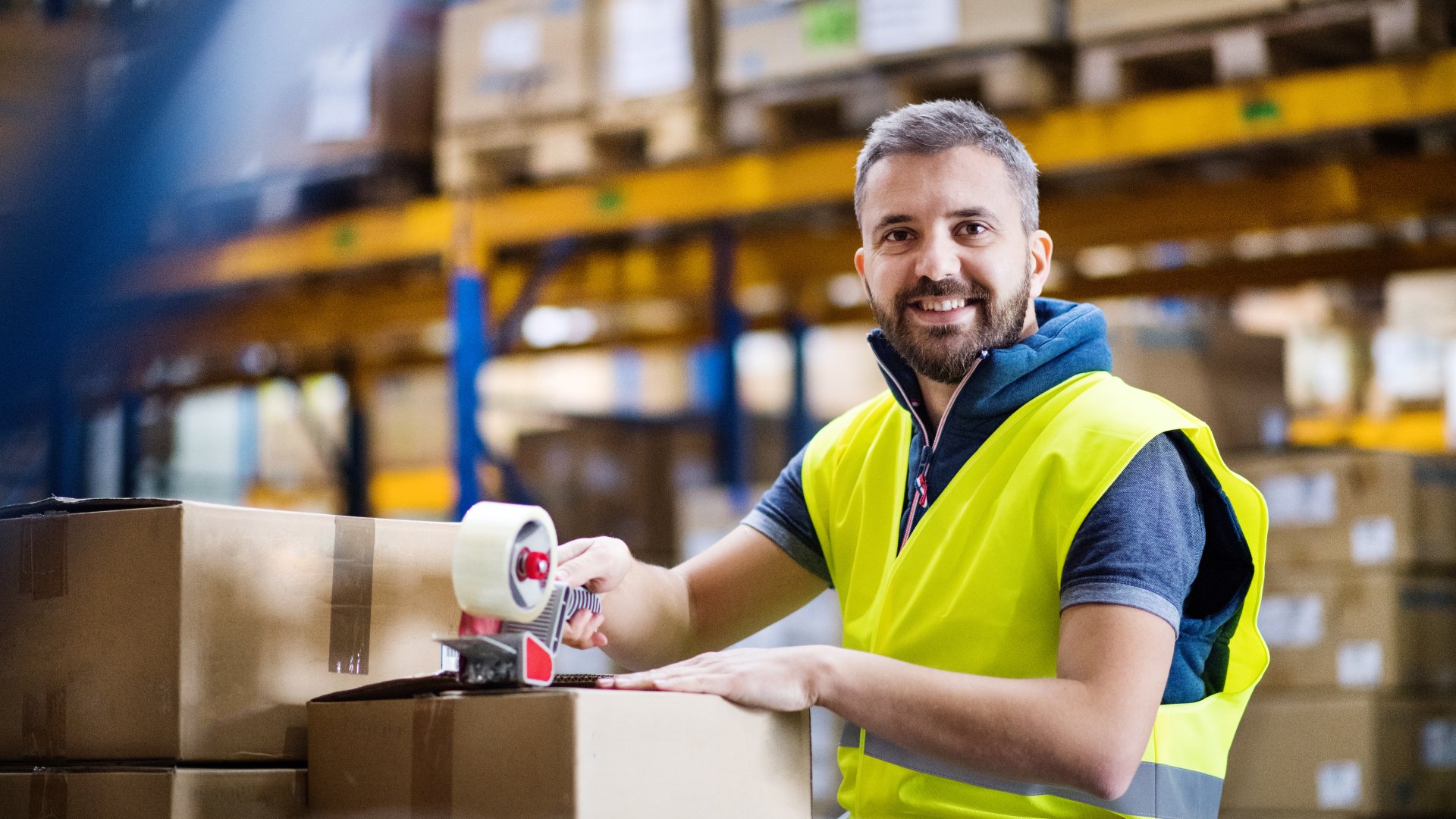 A male warehouse employee is wearing a blue shirt and bright yellow safety vest while he tapes a cardboard box shut.