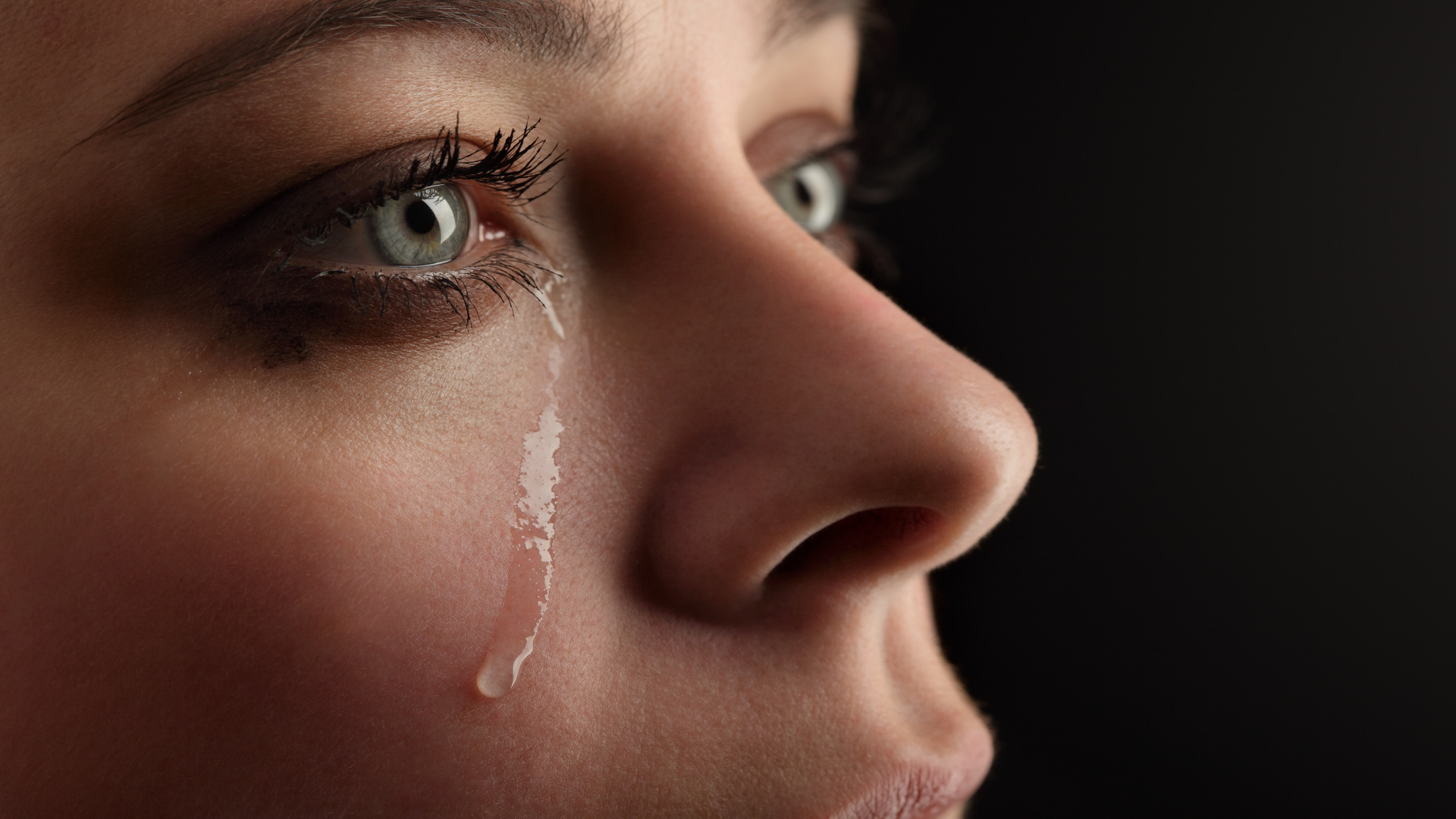 An extreme close-up shows a woman's green eyes and a single tear dripping down her cheek. She is wearing mascara.
