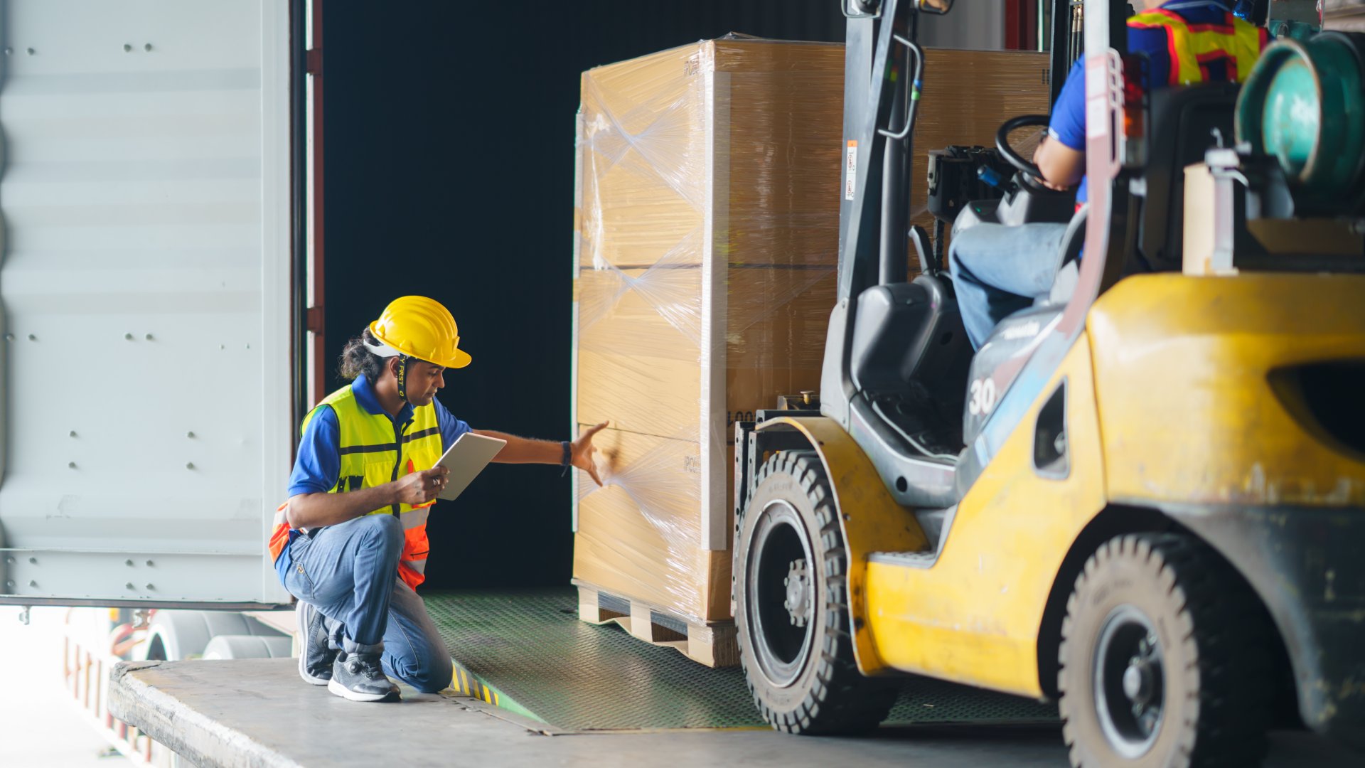 A person kneeling to examine a loaded pallet wrapped in stretch film and resting on the forks of a forklift.