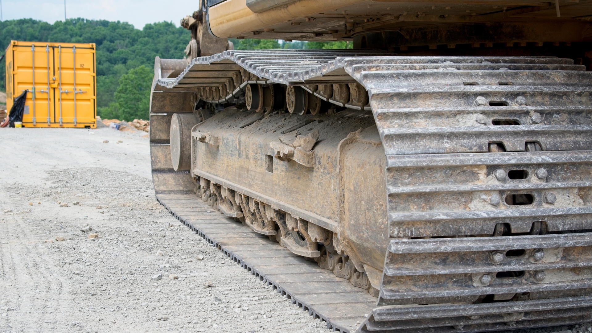 A tracked excavator sits on a jobsite. A yellow shipping container and a row of trees are in the background.