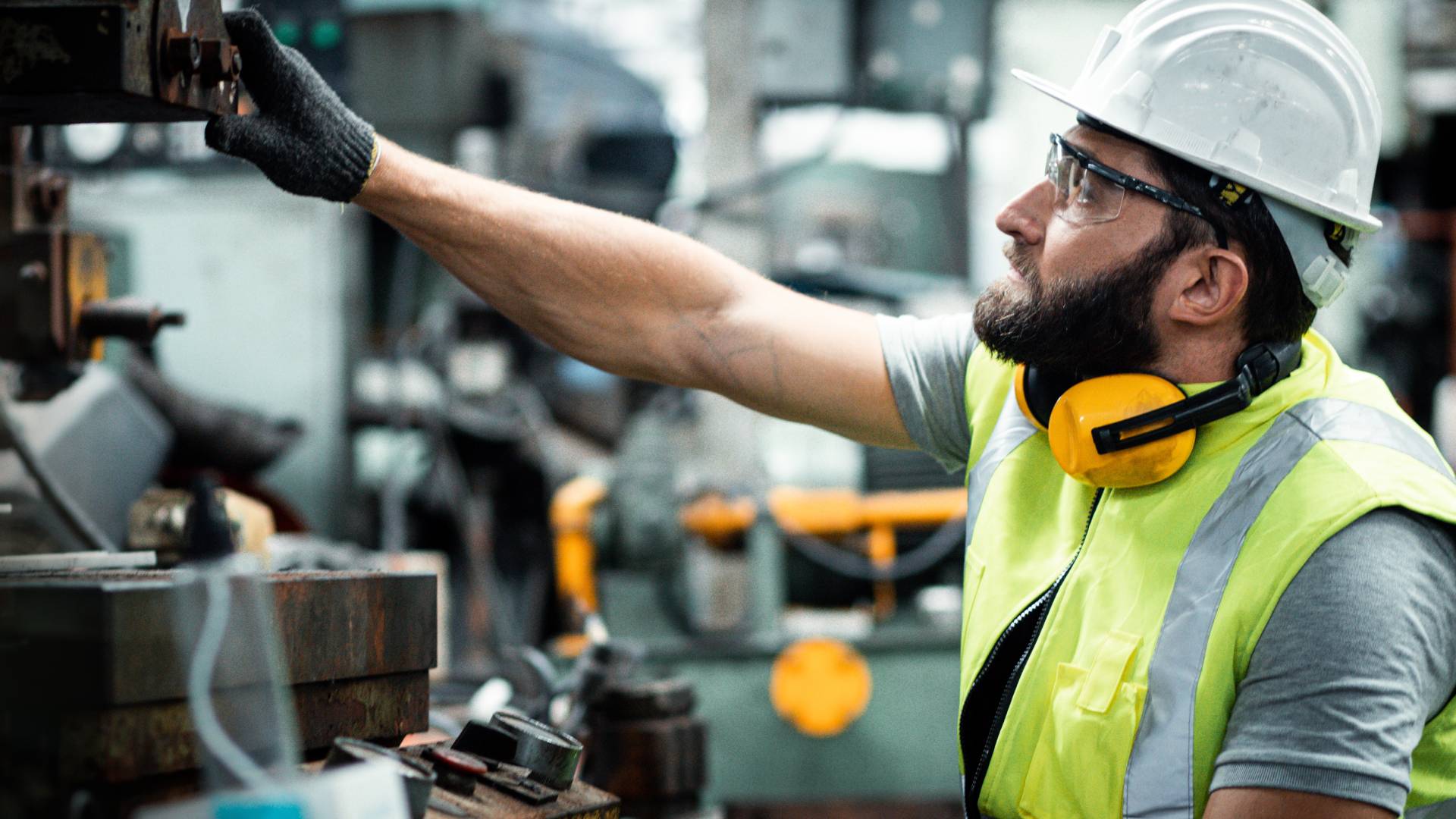 A man wearing a white helmet and other safety gear, working in an industrial warehouse, and checking up on the machinery.