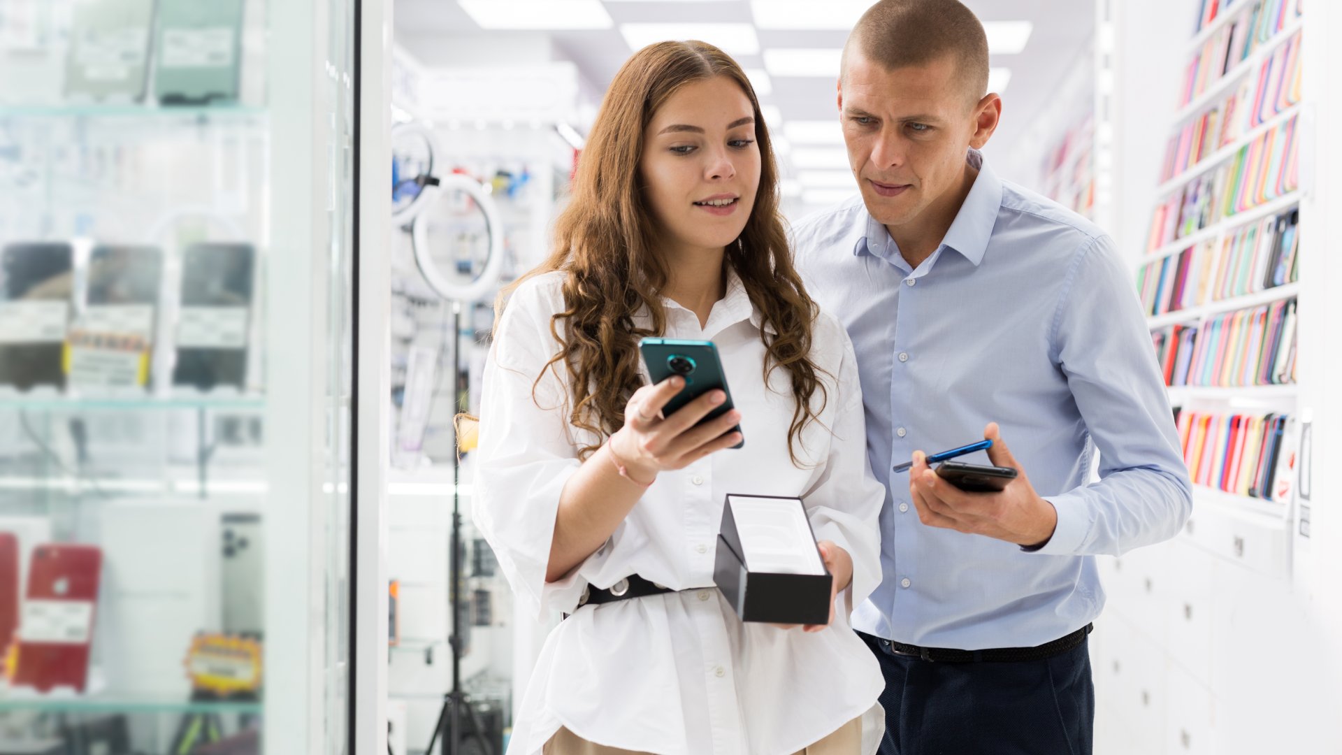 A couple stands in a store, trying to decide which smartphones they should buy. They are looking at three phones.