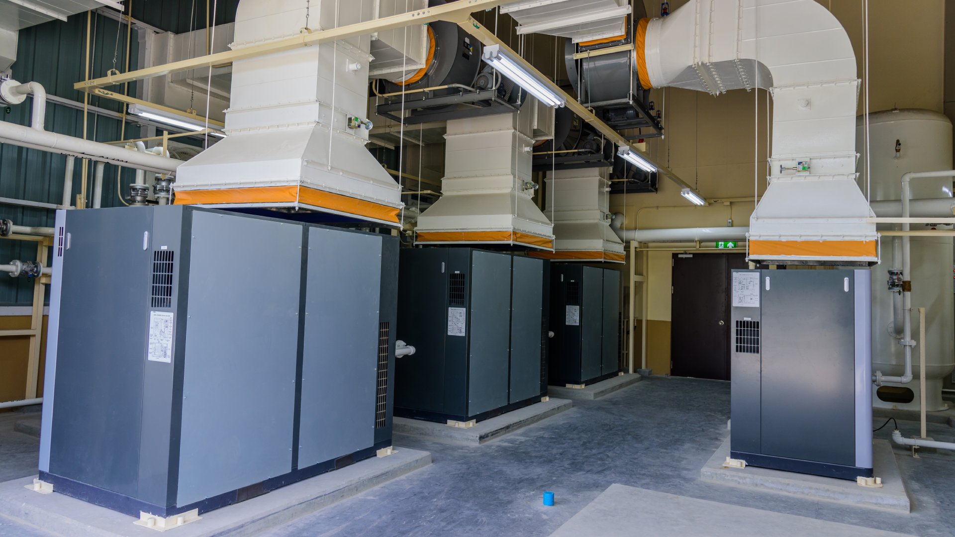 A room with dark gray floors contains large gray pipes coming out of large navy blue industrial air compressors.