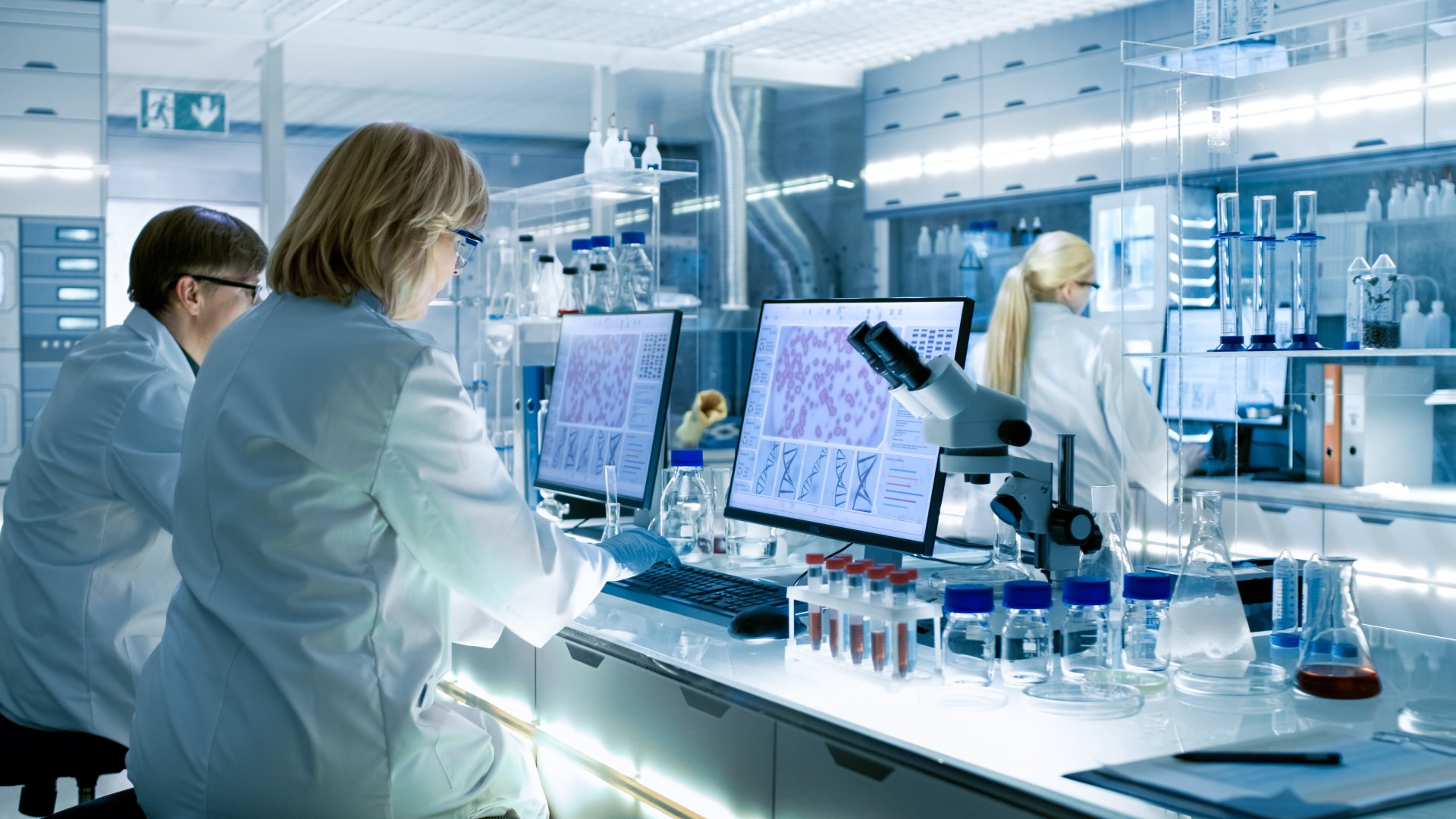 Three scientists in white coats are working in a modern science lab using computers to magnify their findings.