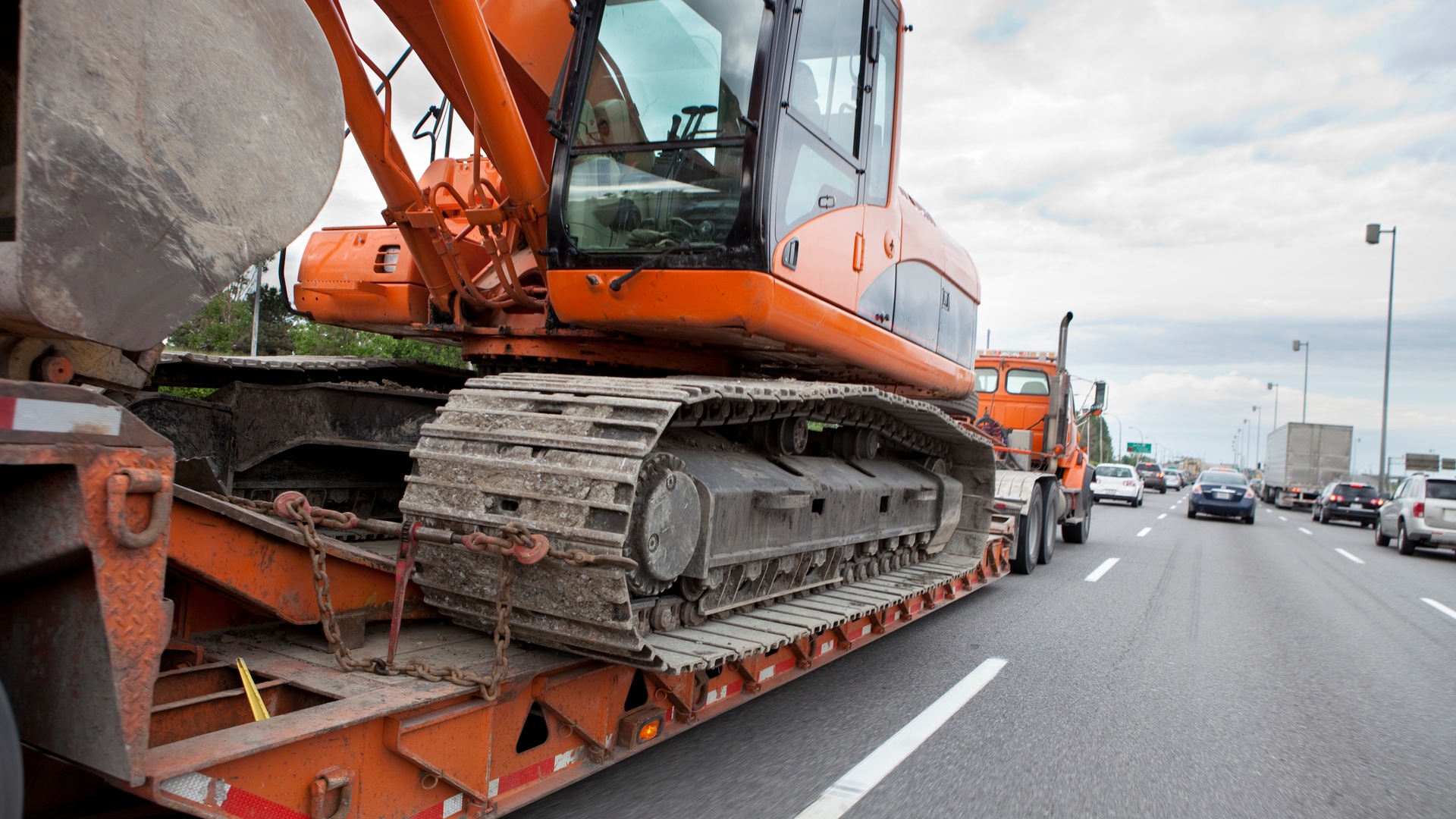 A full-sized orange truck driving down a busy highway. On its back trailer is a large orange excavator crane.