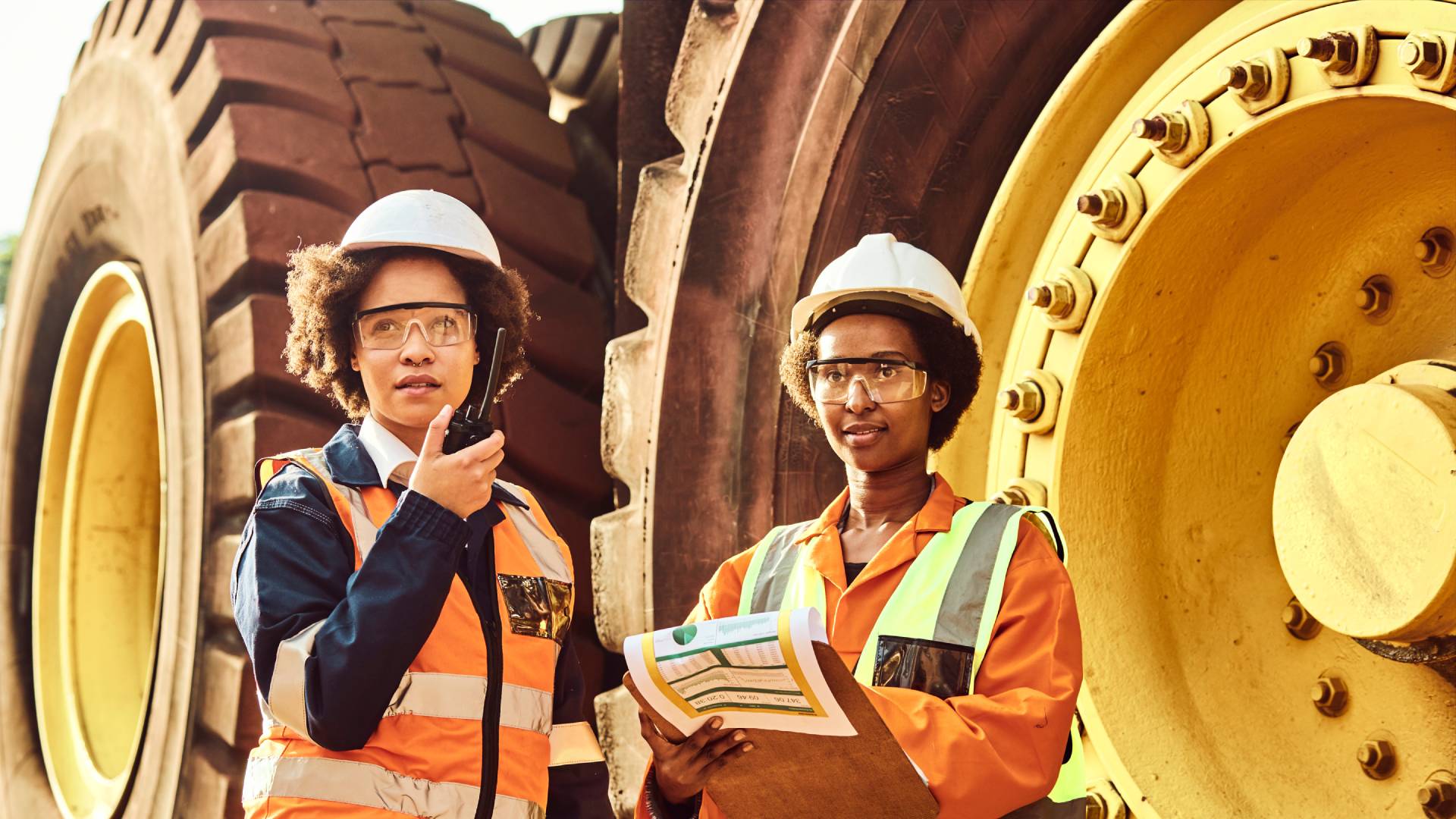 Two female mine workers wearing hi-vis vests and hard hats, surveying mining equipment on a site and writing on a clipboard.