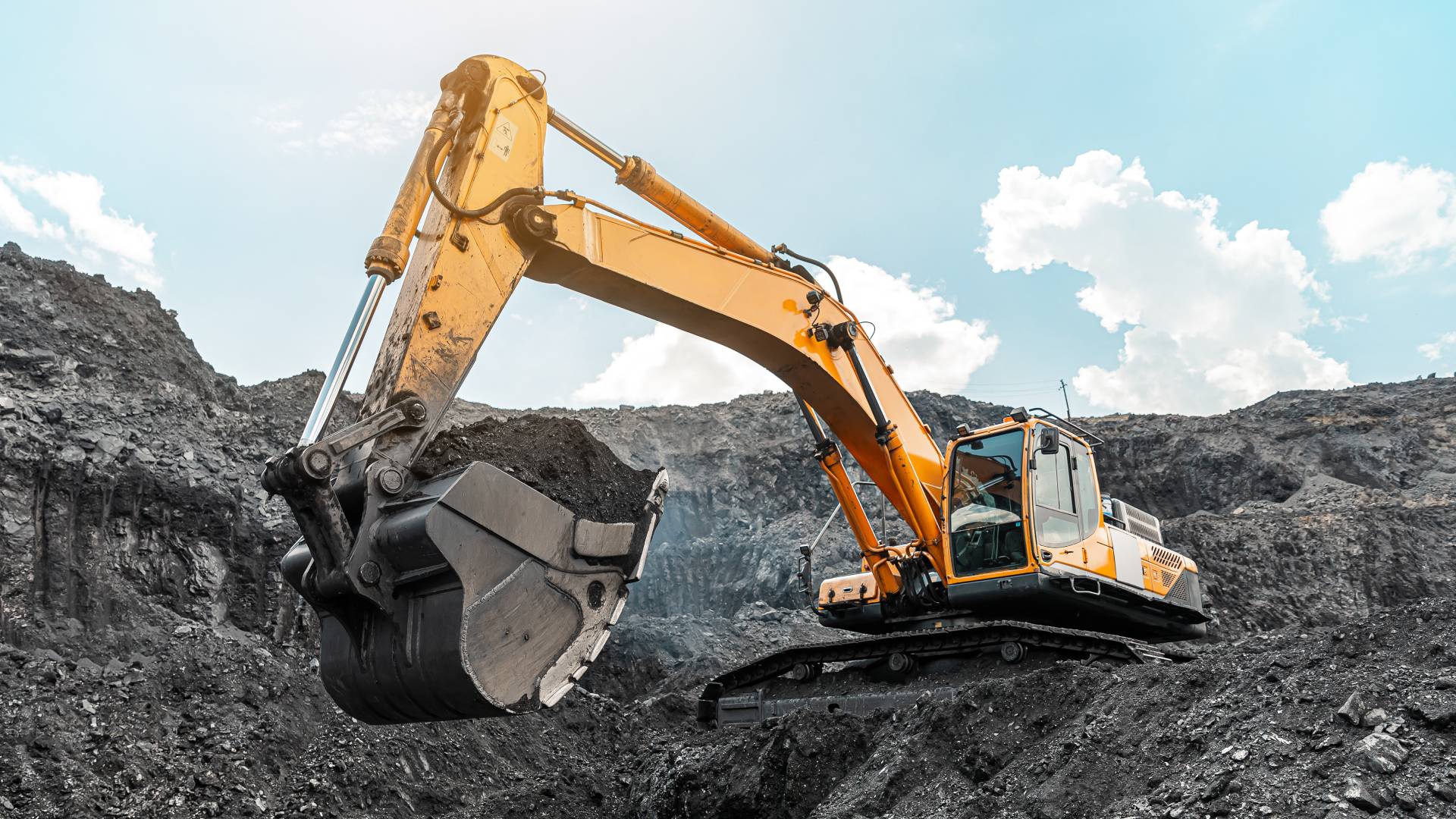 A large excavator sits on top of a hill in a mining quarry, shoveling large amounts of mined material.