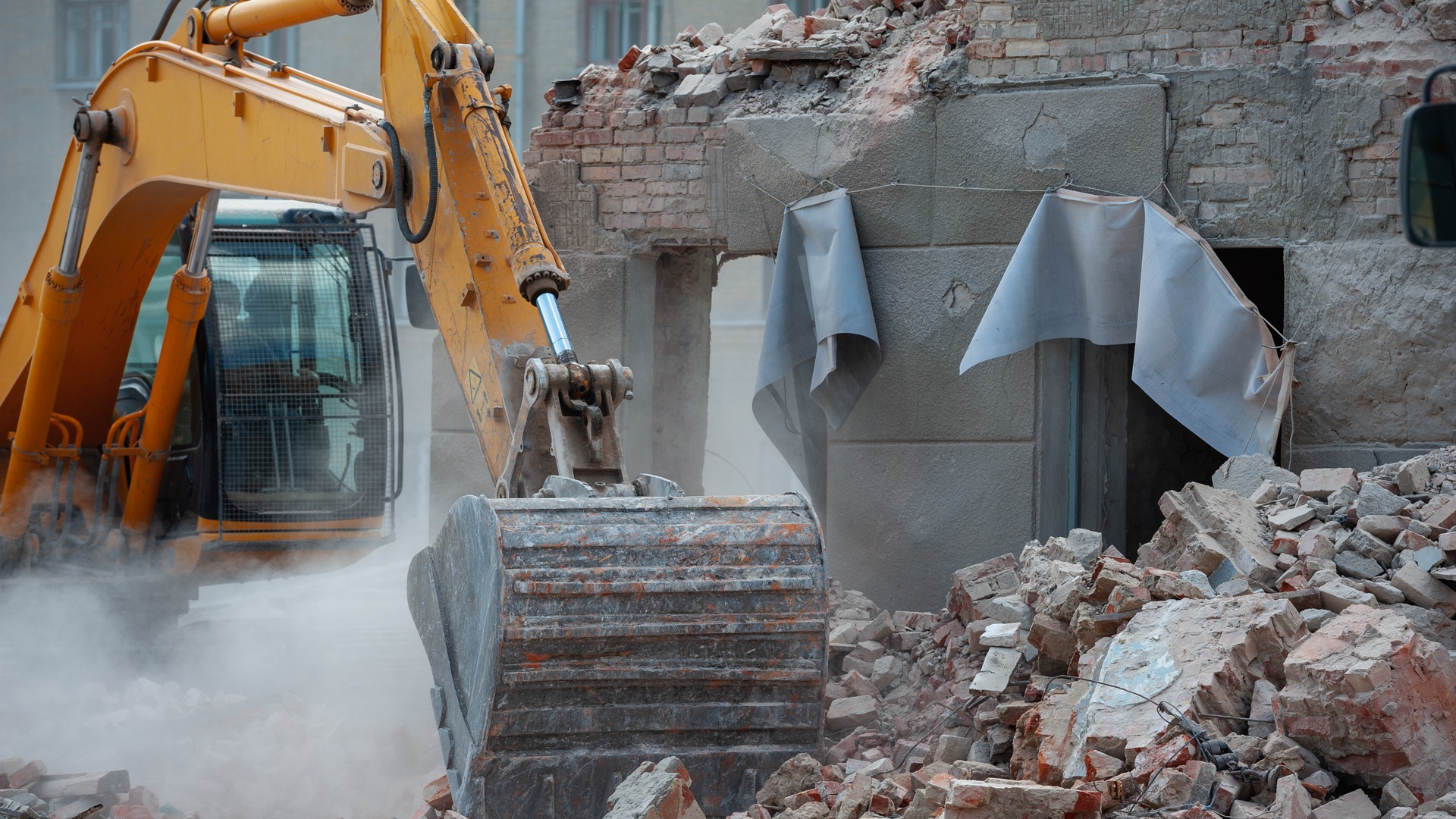 A yellow excavator is used to break down a building made of bricks, its toothed scoop resting on the pile of debris.
