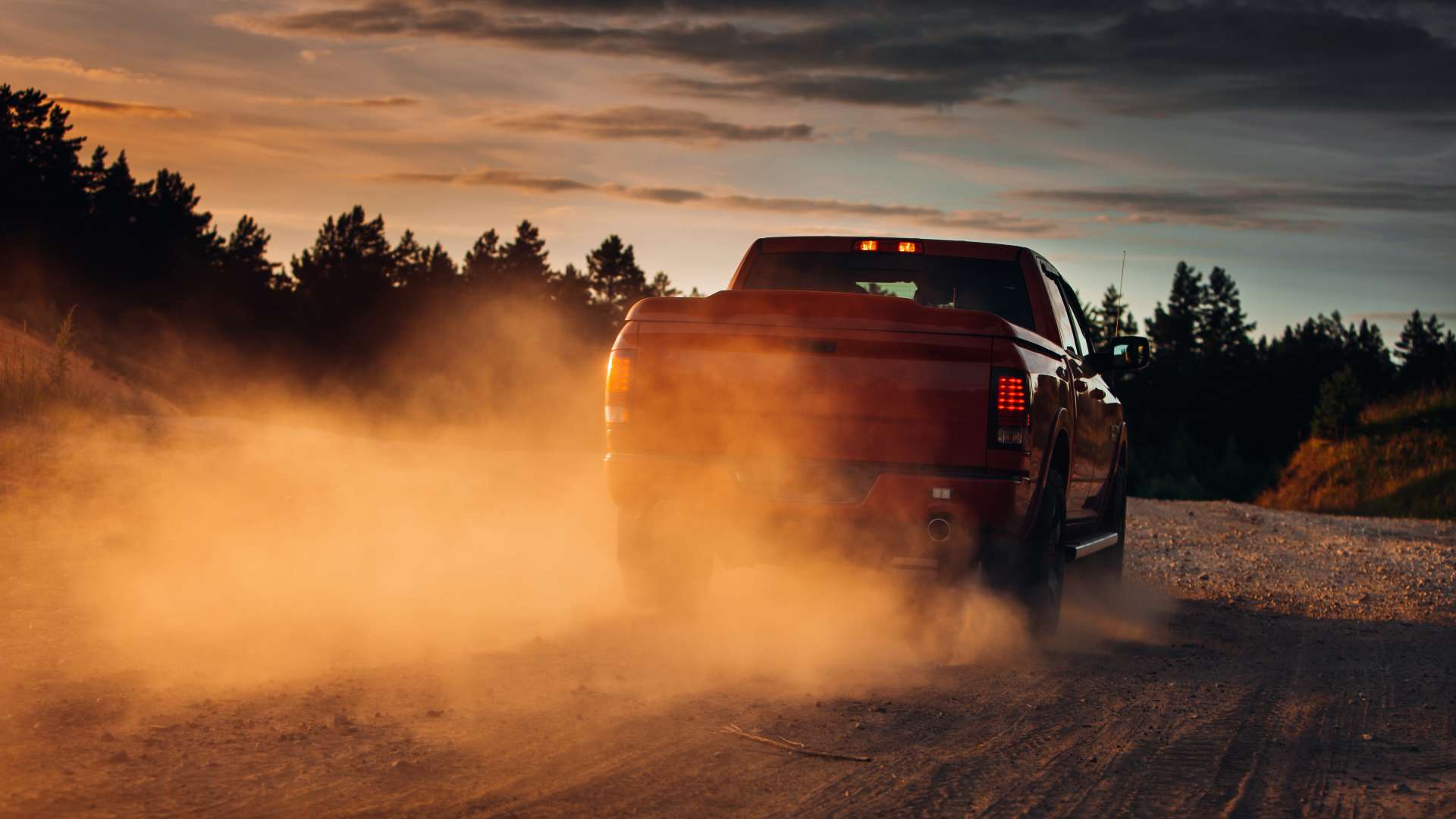 A red pickup truck driving along a gravel path during sunset. The truck's motion is kicking up a great deal of dust.