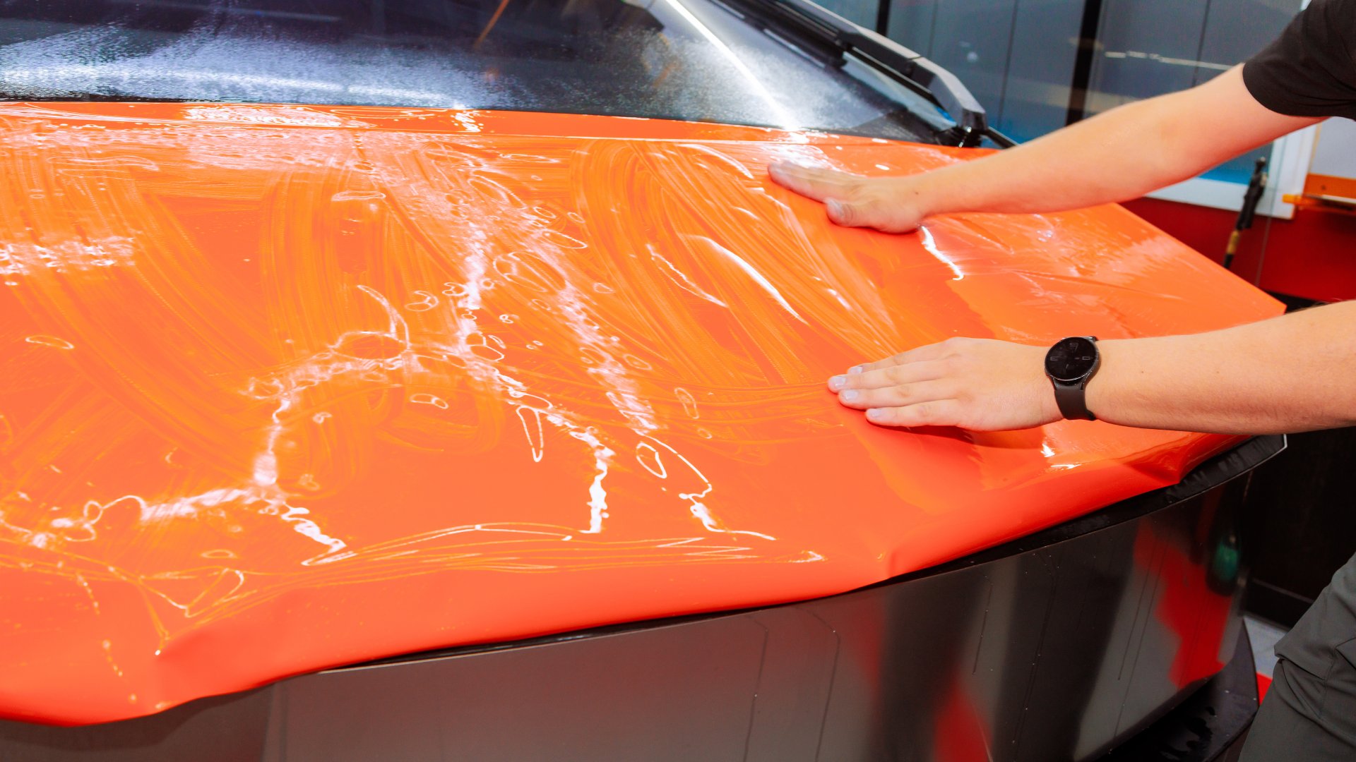 A person with a black watch on their left arm uses both hands to smooth out an orange vinyl wrap over the hood of a car.