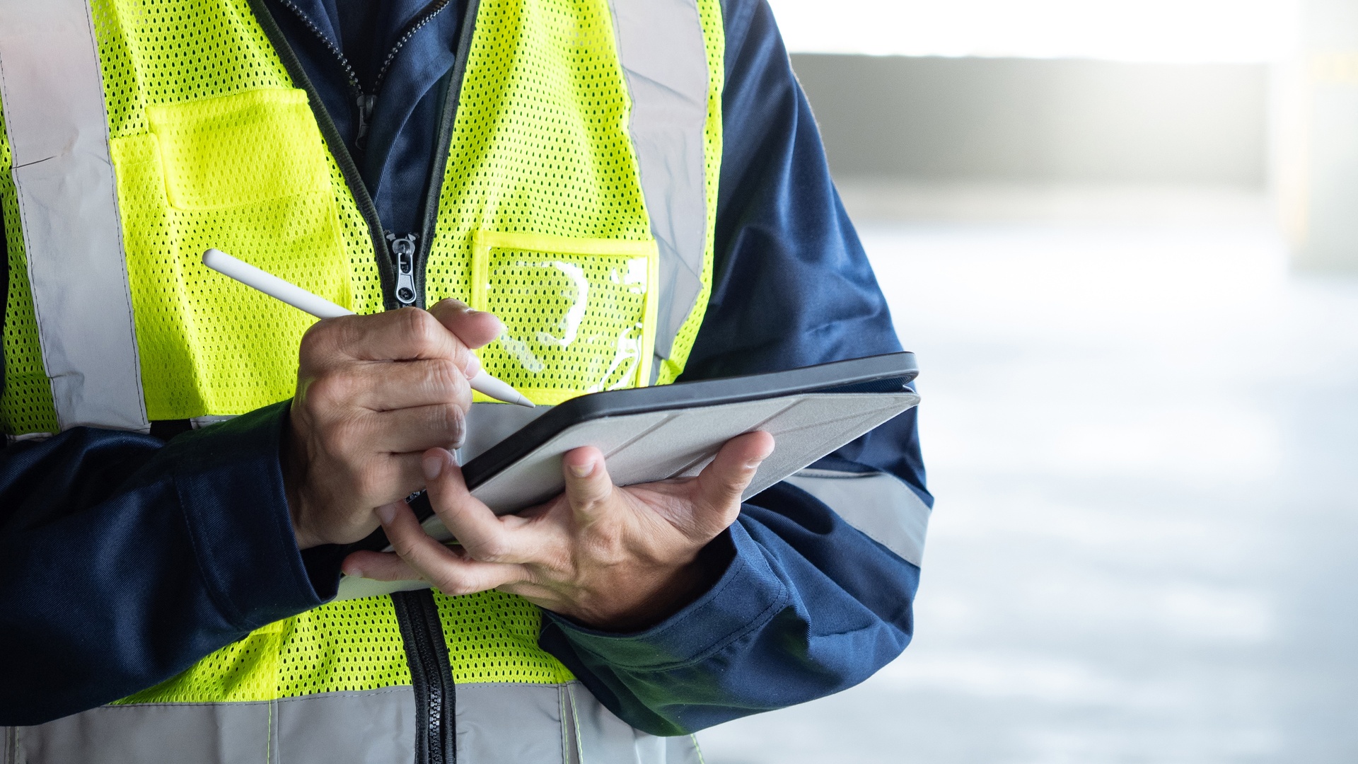 A worker is wearing a high-visibility vest. He's holding a tablet in one hand and using a stylus in the other.