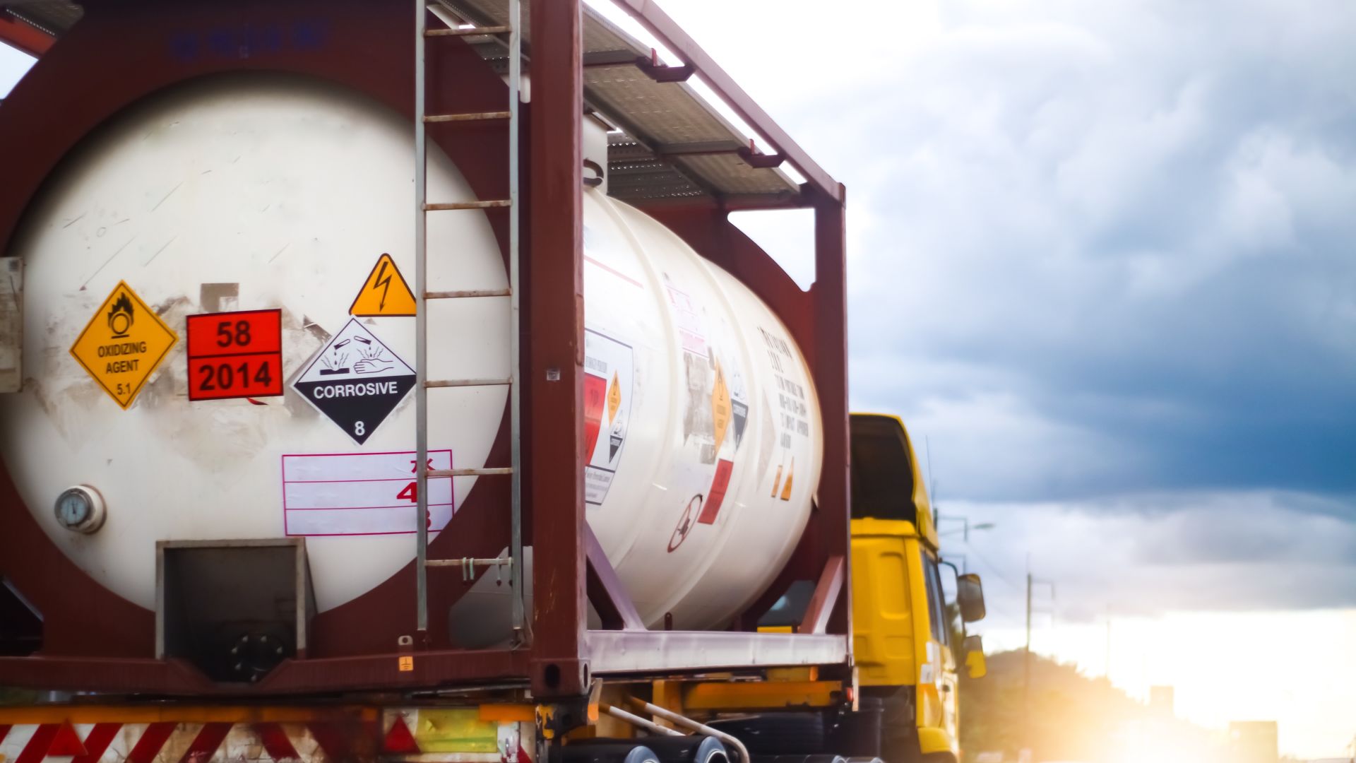 A truck with a yellow cab transporting a white tank of corrosive chemicals labeled with several safety stickers.
