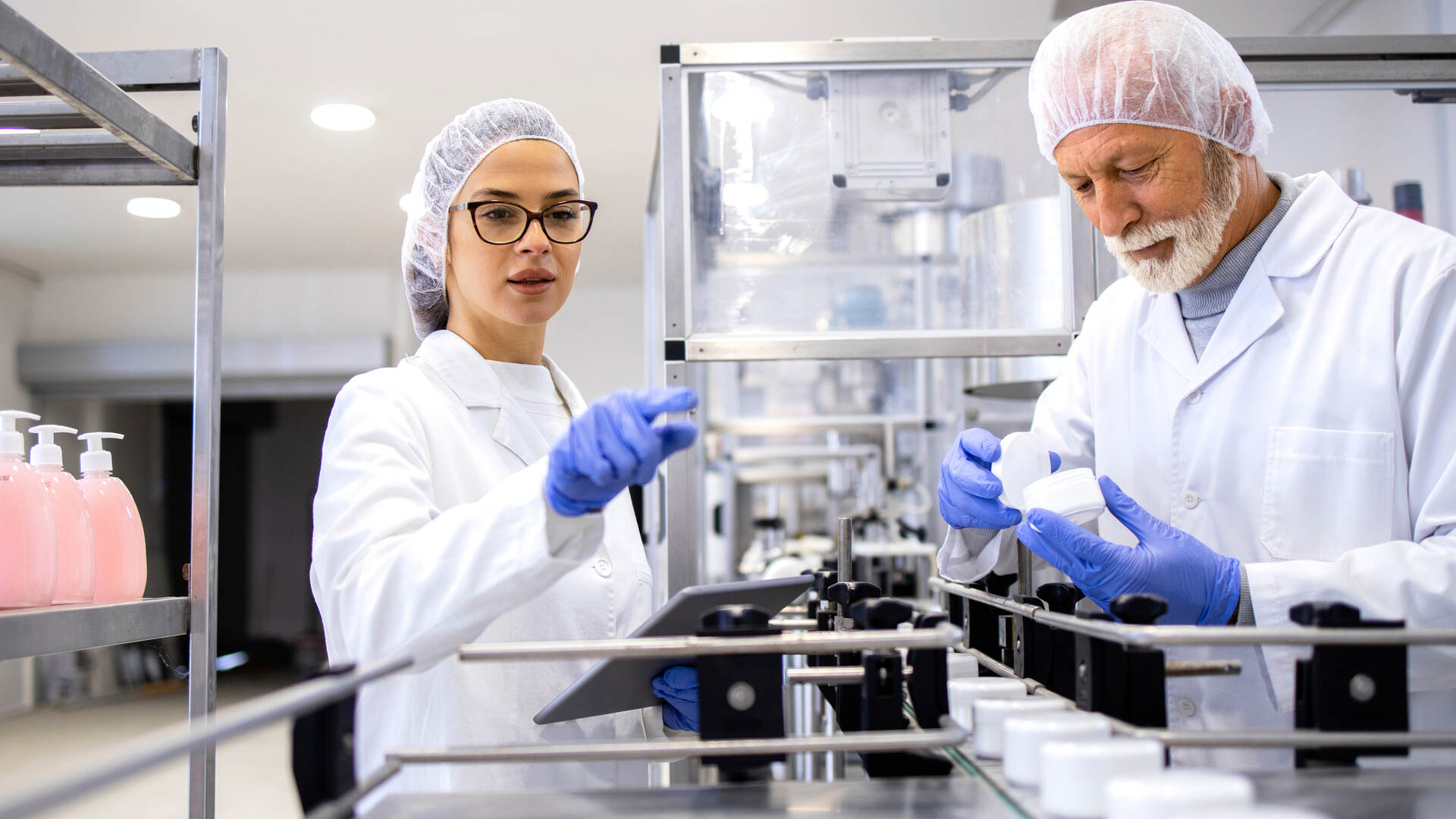 Two workers wearing latex gloves, hair nets, and lab coats are working on a line with white jars on a conveyor.