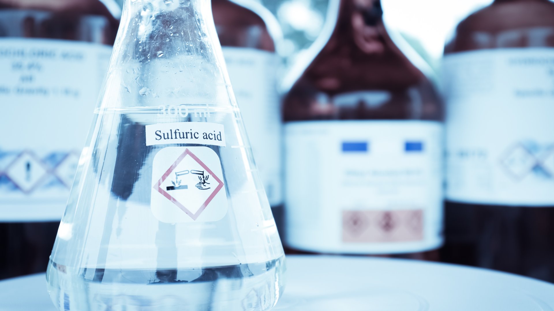 A clear beaker labeled "SULFURIC ACID" sits on a table in a laboratory. Brown beakers sit behind it.