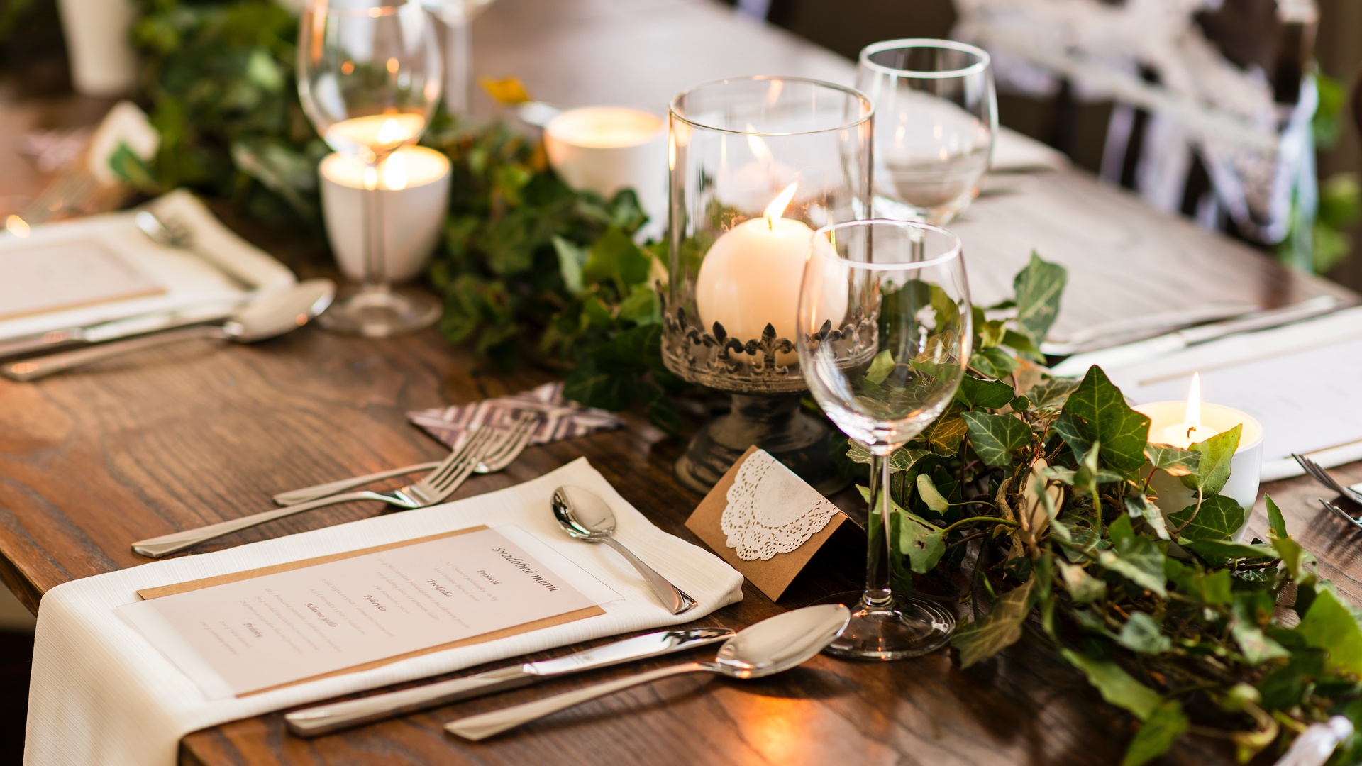 A close-up of a table setting at a wedding with a menu, wineglasses, candles, greenery, napkins, and silverware.