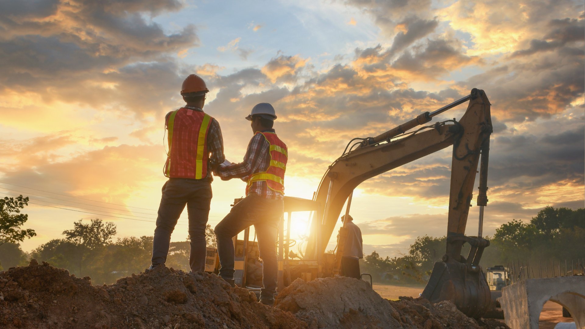 Two workers wearing hard hats standing in front of a large excavator on a construction site as the sun sets.