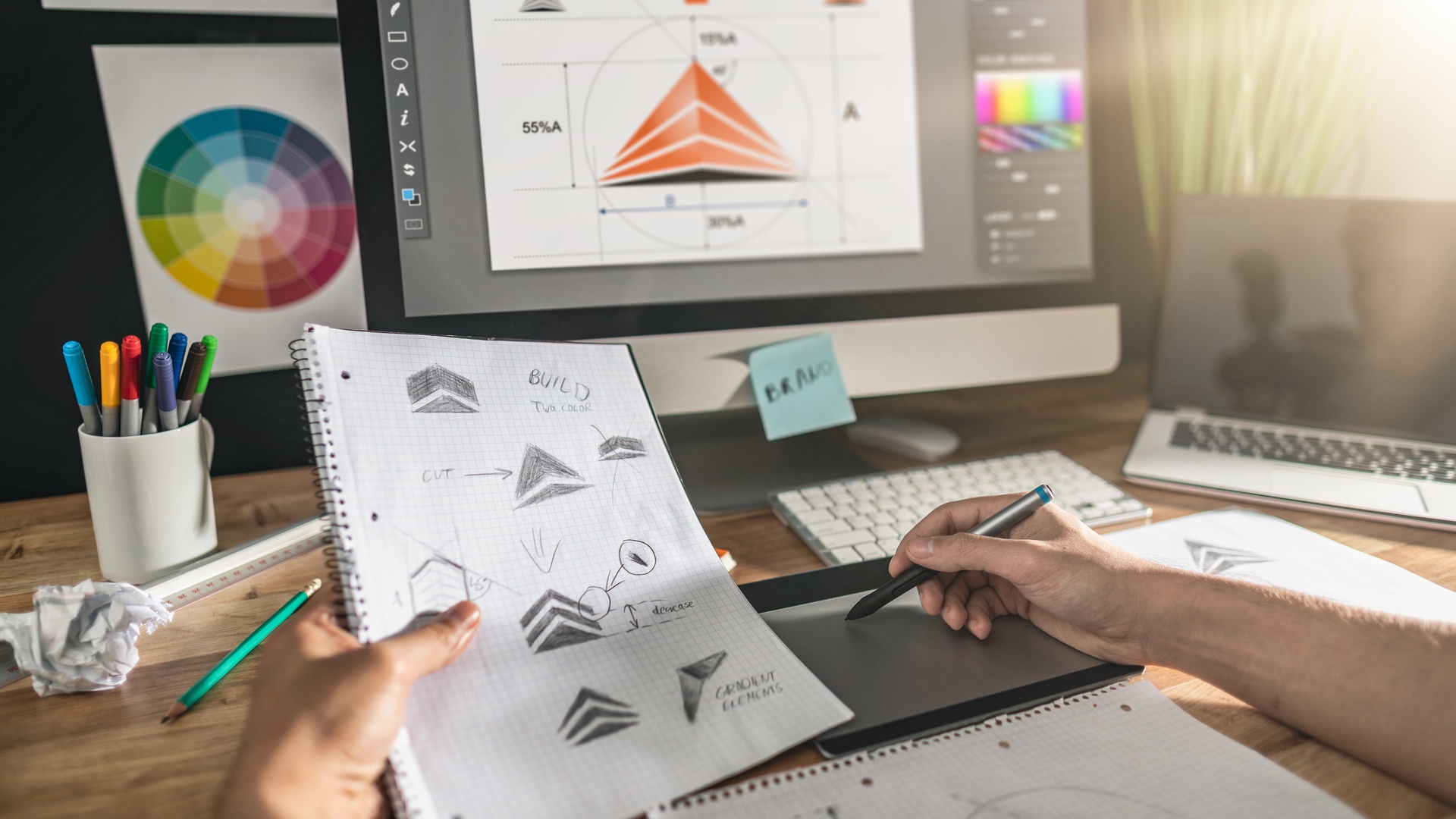 A close-up of a person sitting at a desk and working on a laptop that shows a logo. They hold a sketchbook in one hand with sketches of the same logo.