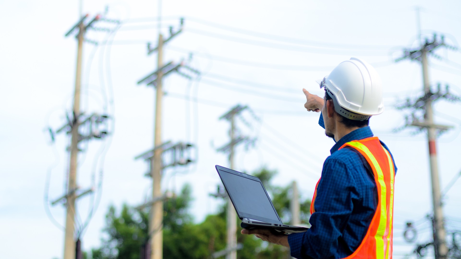 A utility worker wearing PPE hat and vest and holding a laptop computer points at power lines in the distance.