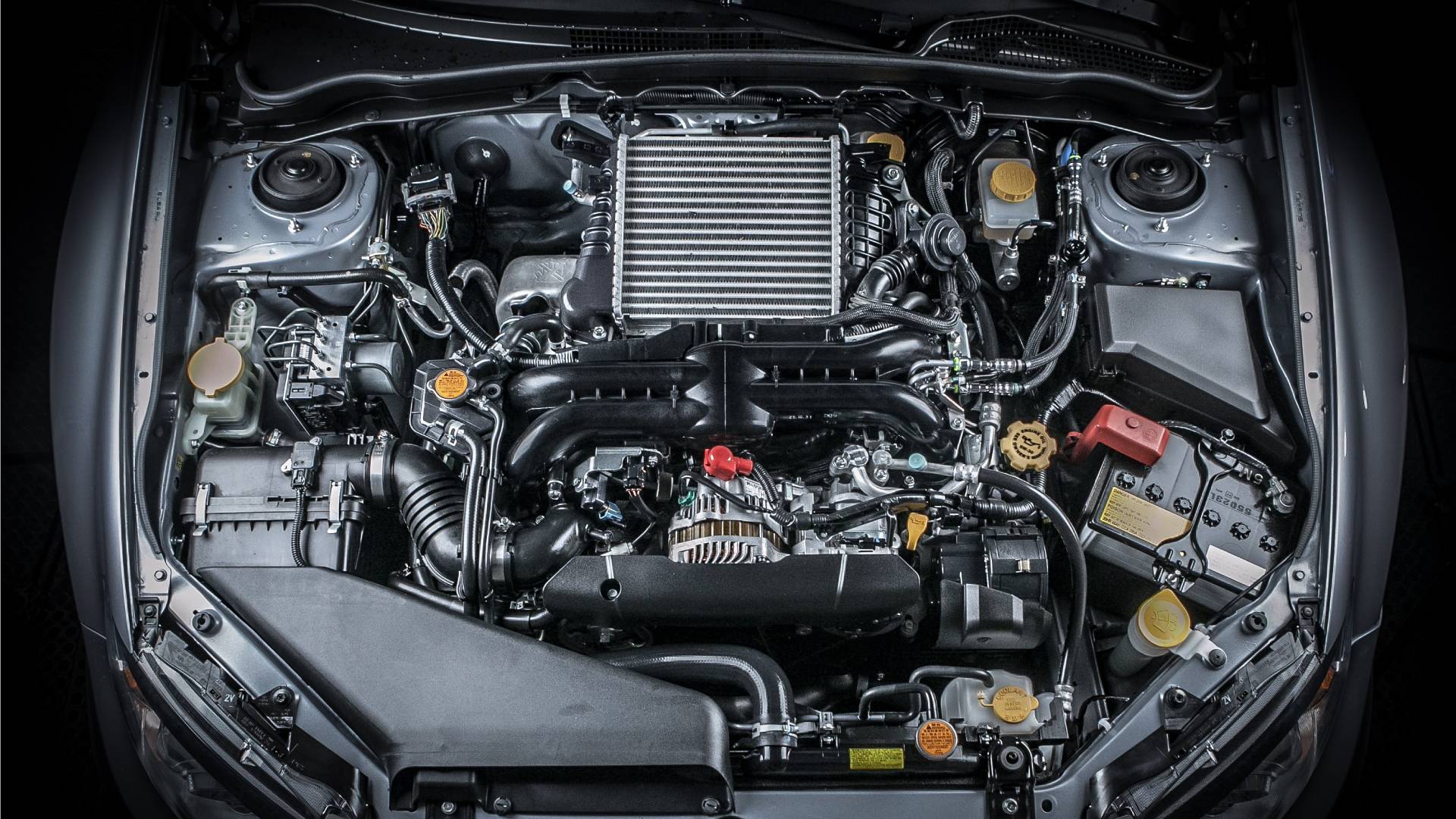 A car has their hood up and there are tons of different components, tubes, and metal parts next to the engine in the middle.
