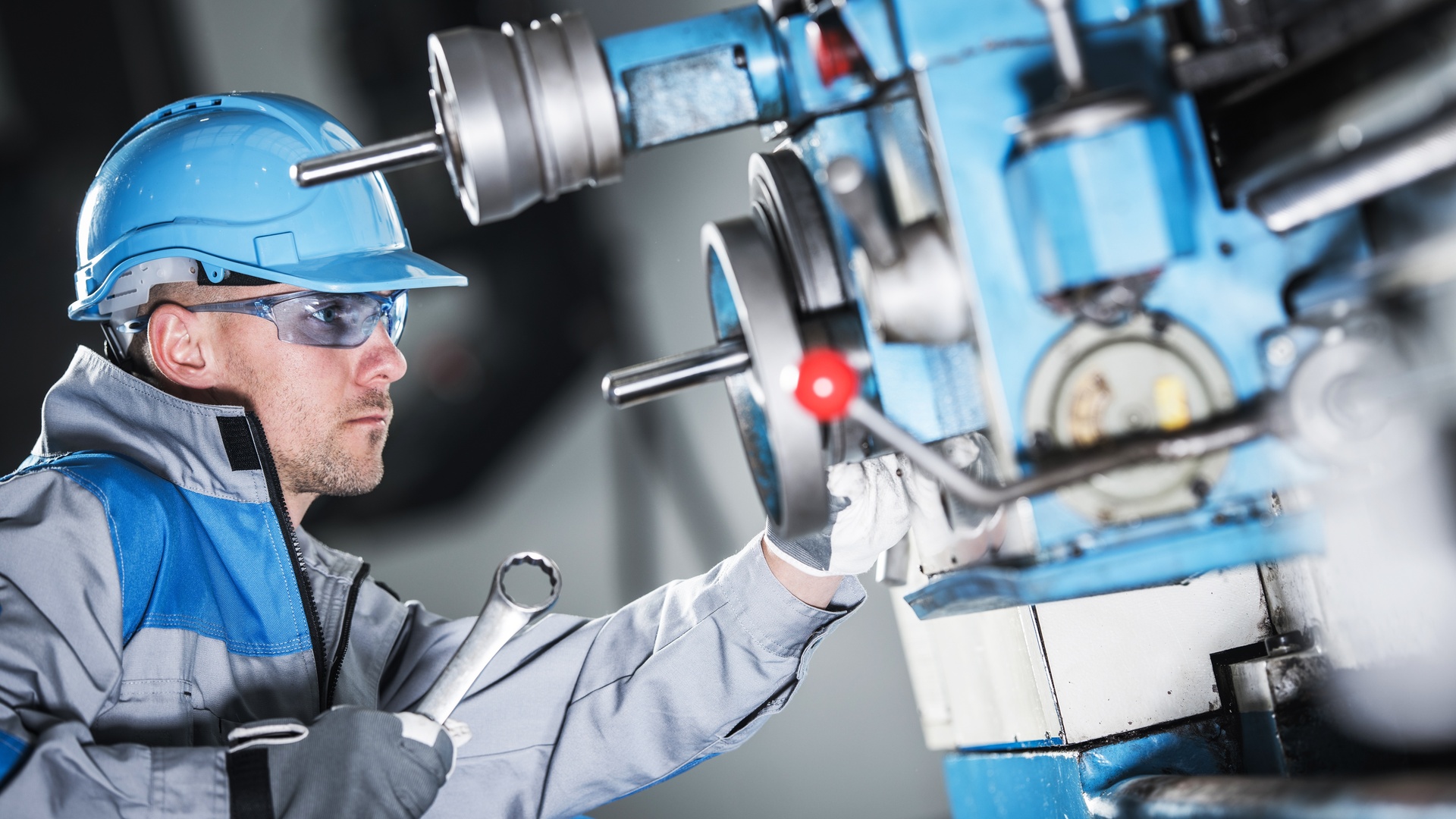 A maintenance man wearing a light blue helmet holds a wrench to a machine, the machine's blue color matching his helmet.