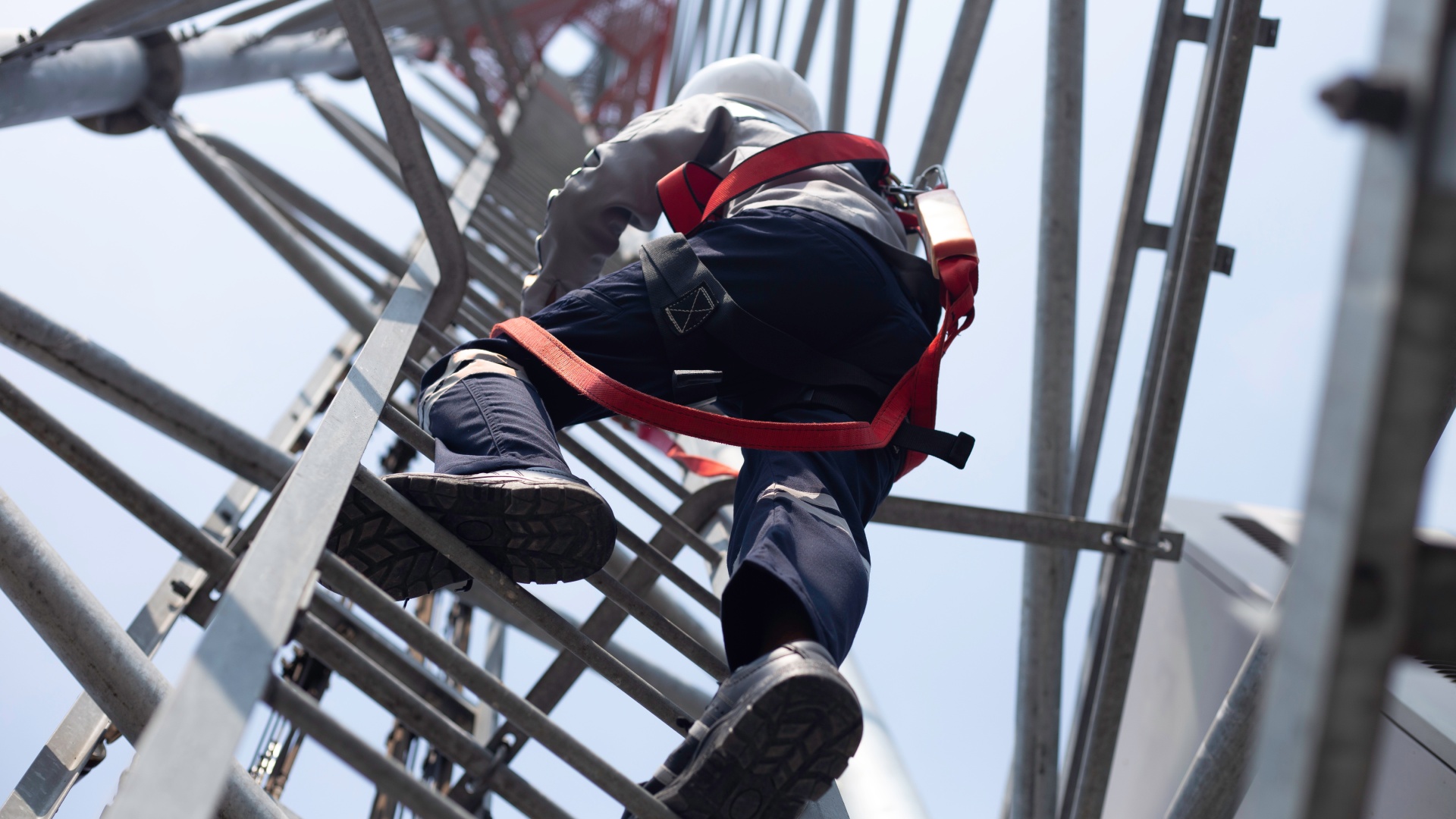 An engineer in a hard hat is climbing up a ladder on a communication tower while wearing a red safety harness.