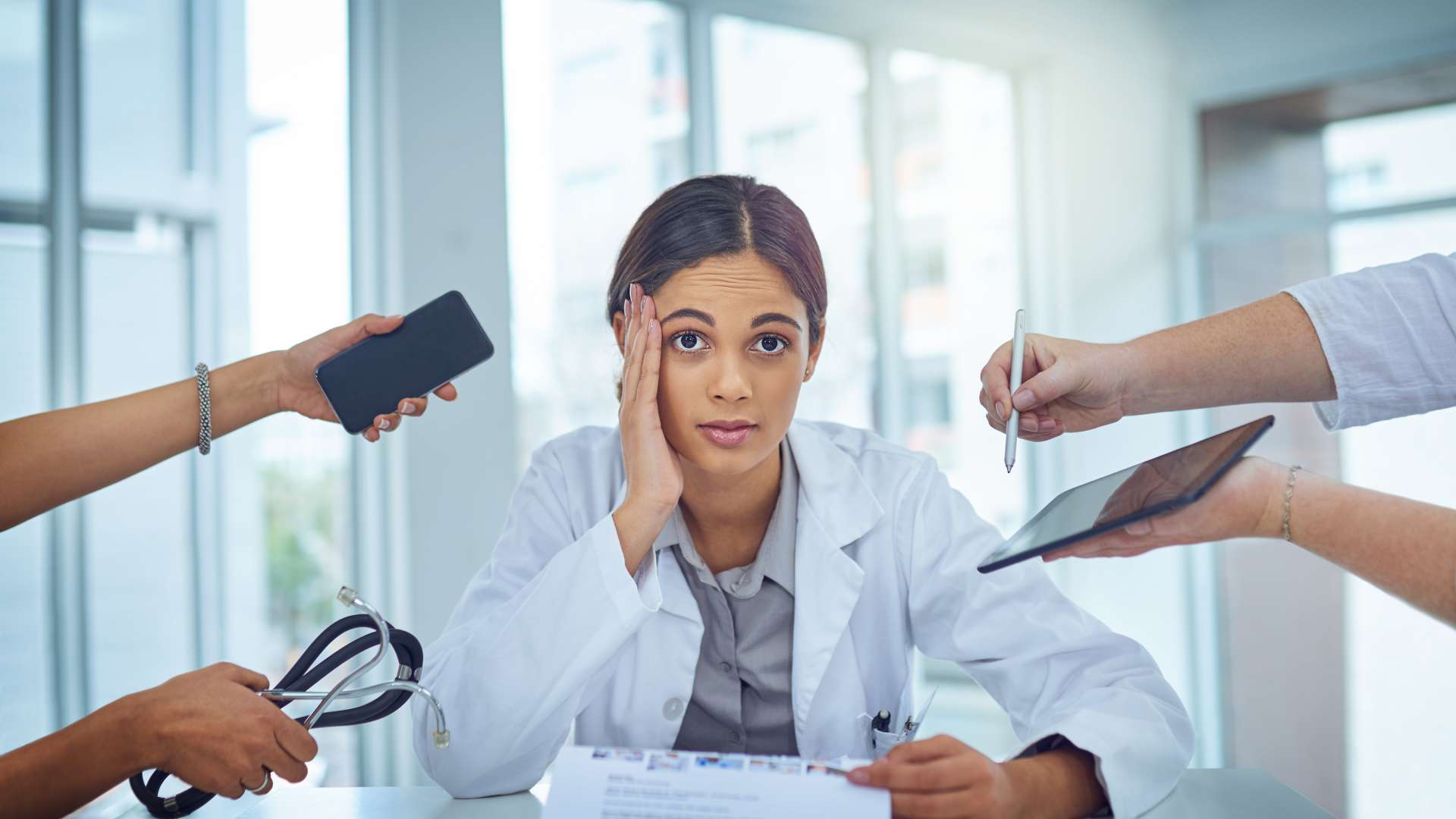 A woman at a desk wearing a white lab coat and holding some forms. Hands holding a stethoscope and smart devices surround her.