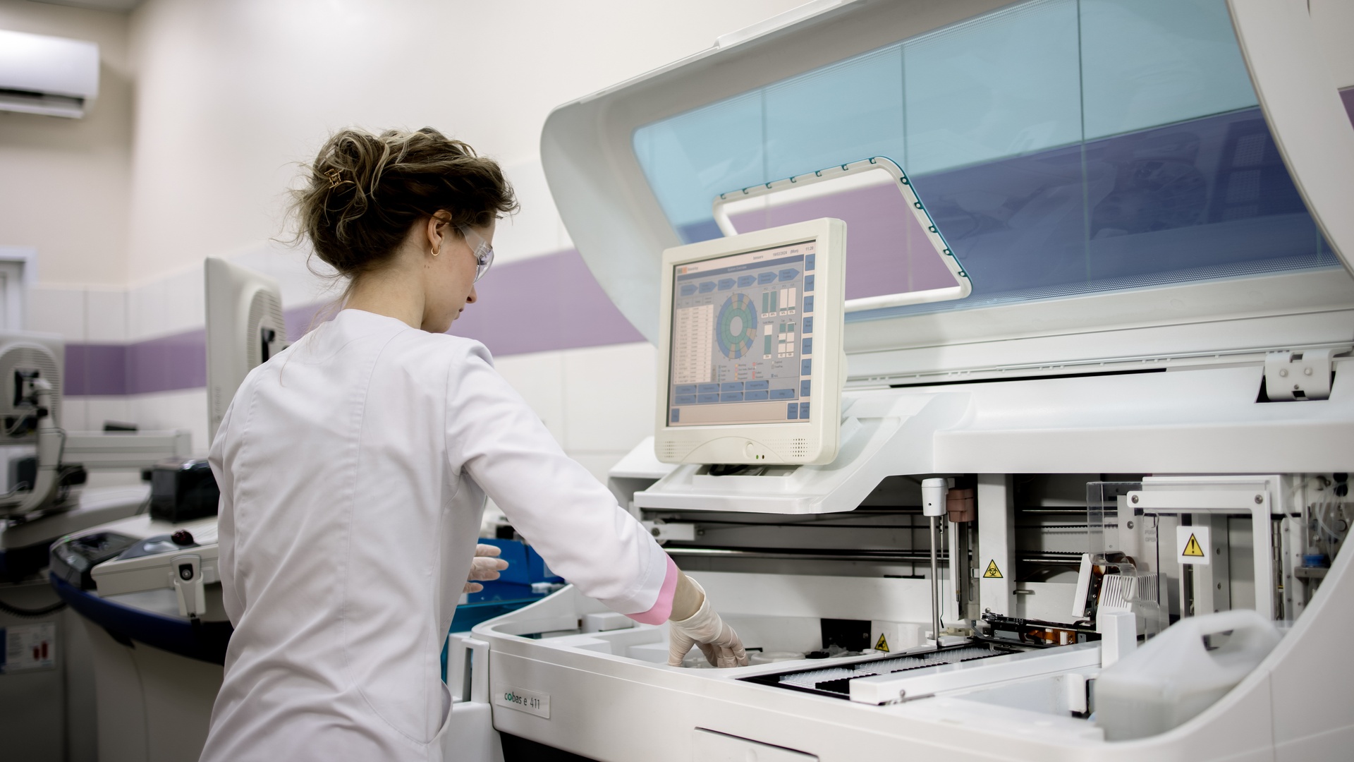 A laboratory professional in a white coat and safety glasses as she tests a medical device in a development machine.