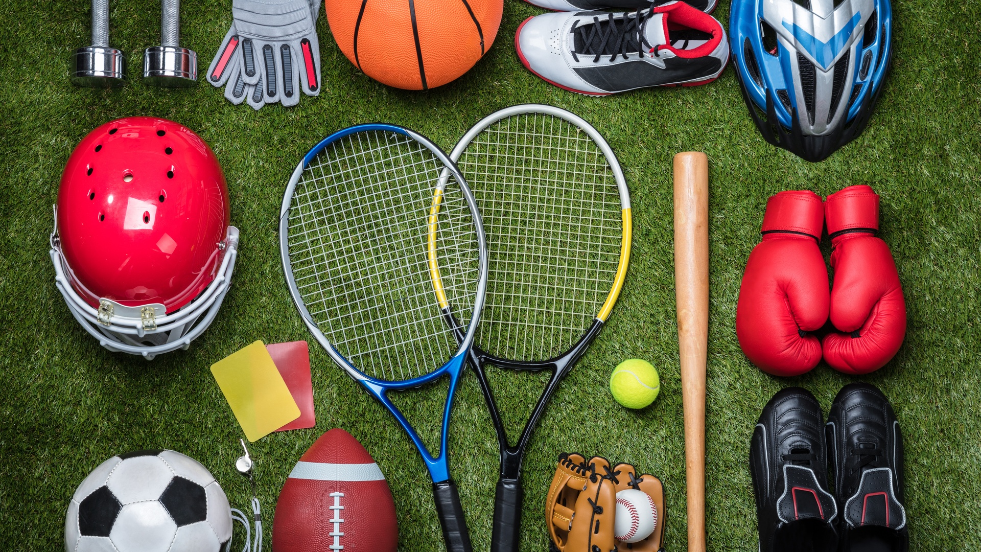 An overhead view of various sporting equipment resting on grass, including balls, rackets, shoes, and helmets.