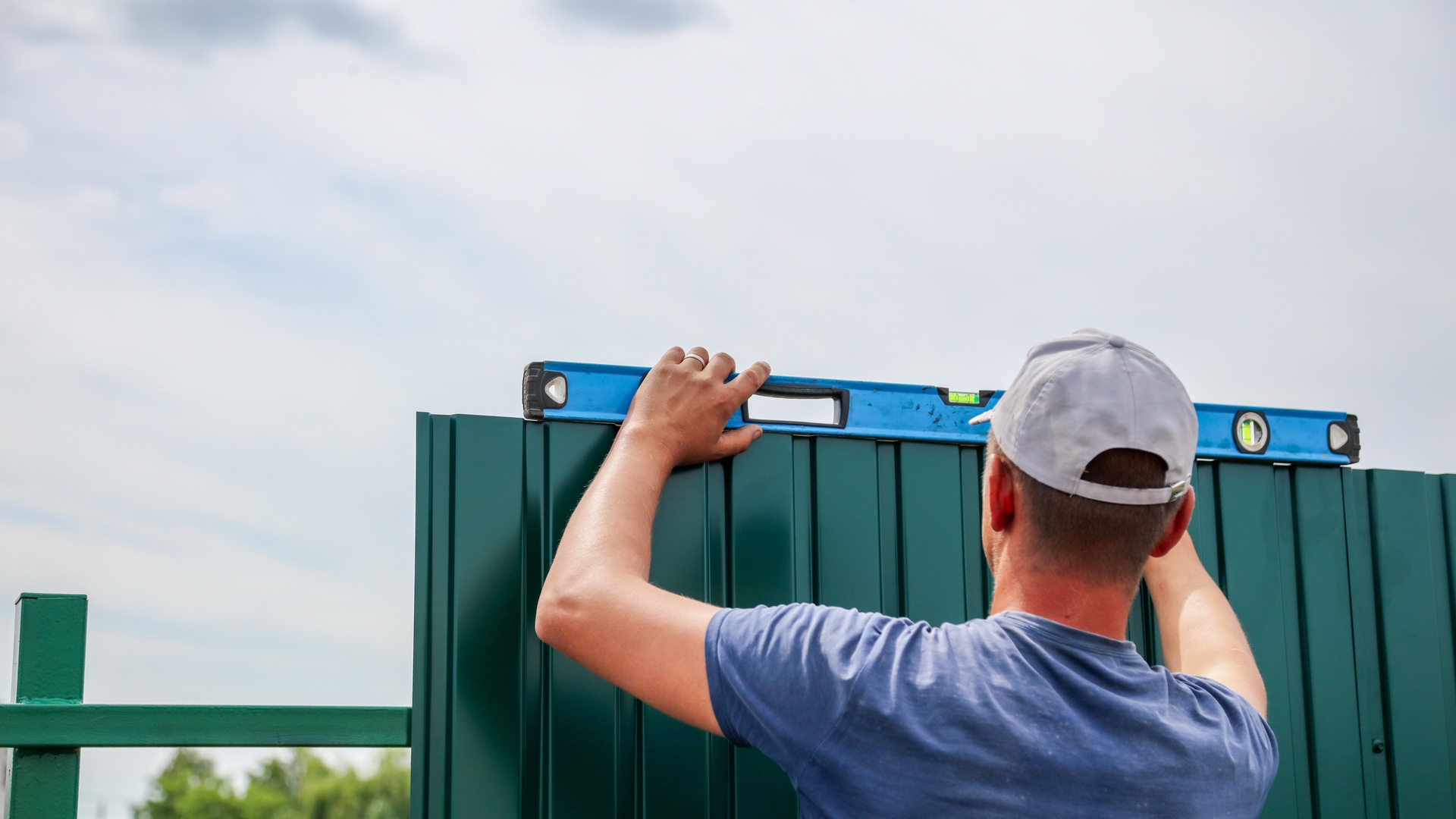 The rear view of a man installing a tall, green fence. He uses a level to check the top of the fence.