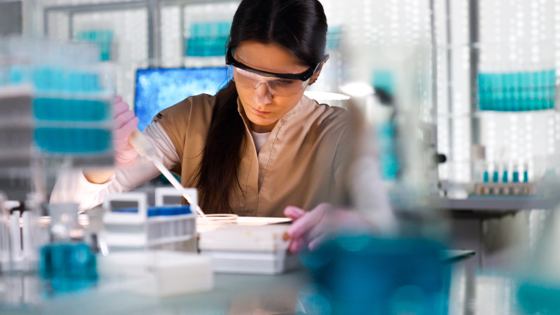 A scientist wearing safety goggles and gloves uses a long, white tool to prepare samples in a laboratory.
