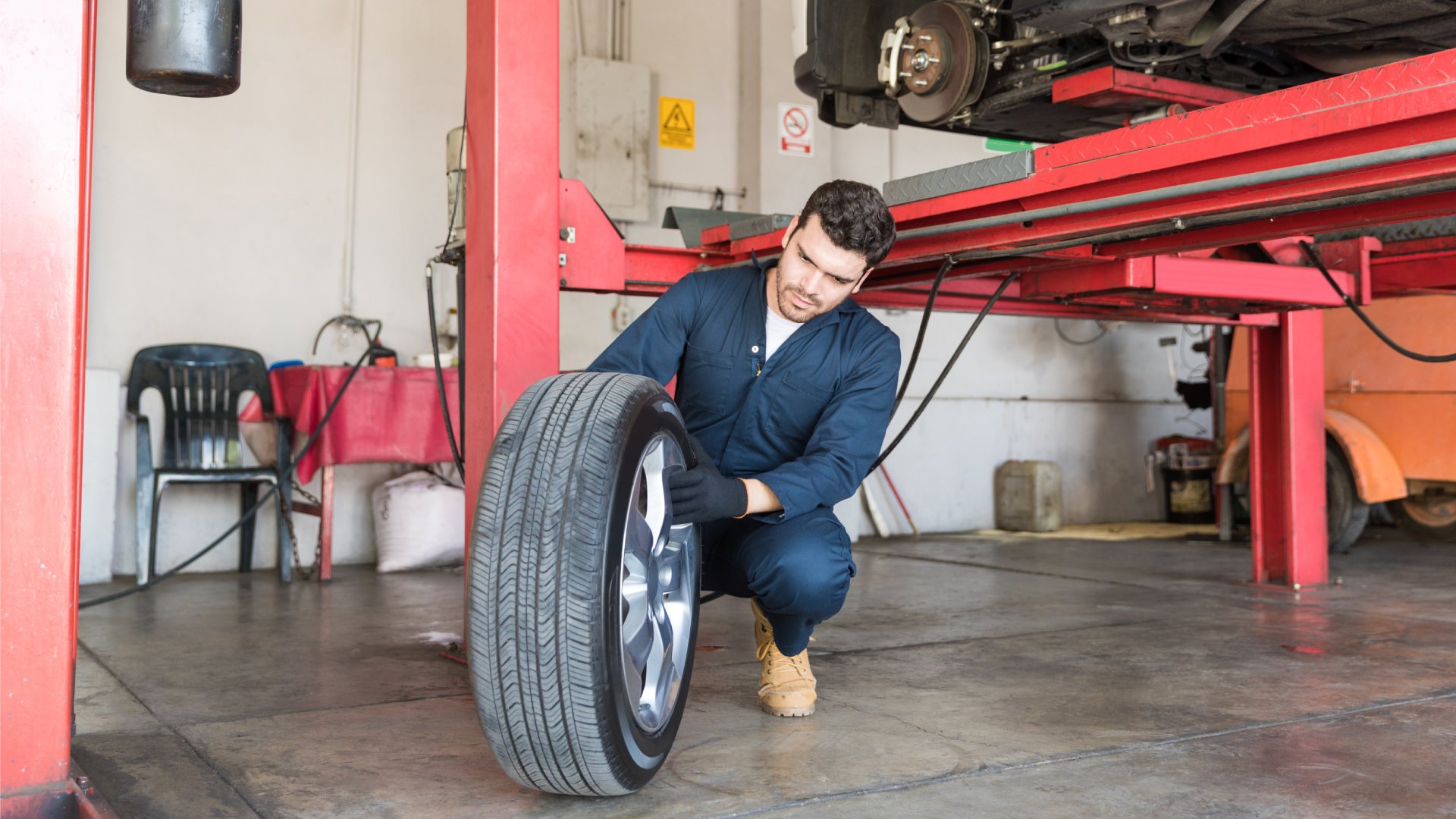 A mechanic in a blue jumpsuit changes a tire on a white car that's sitting on a red four-post auto lift.