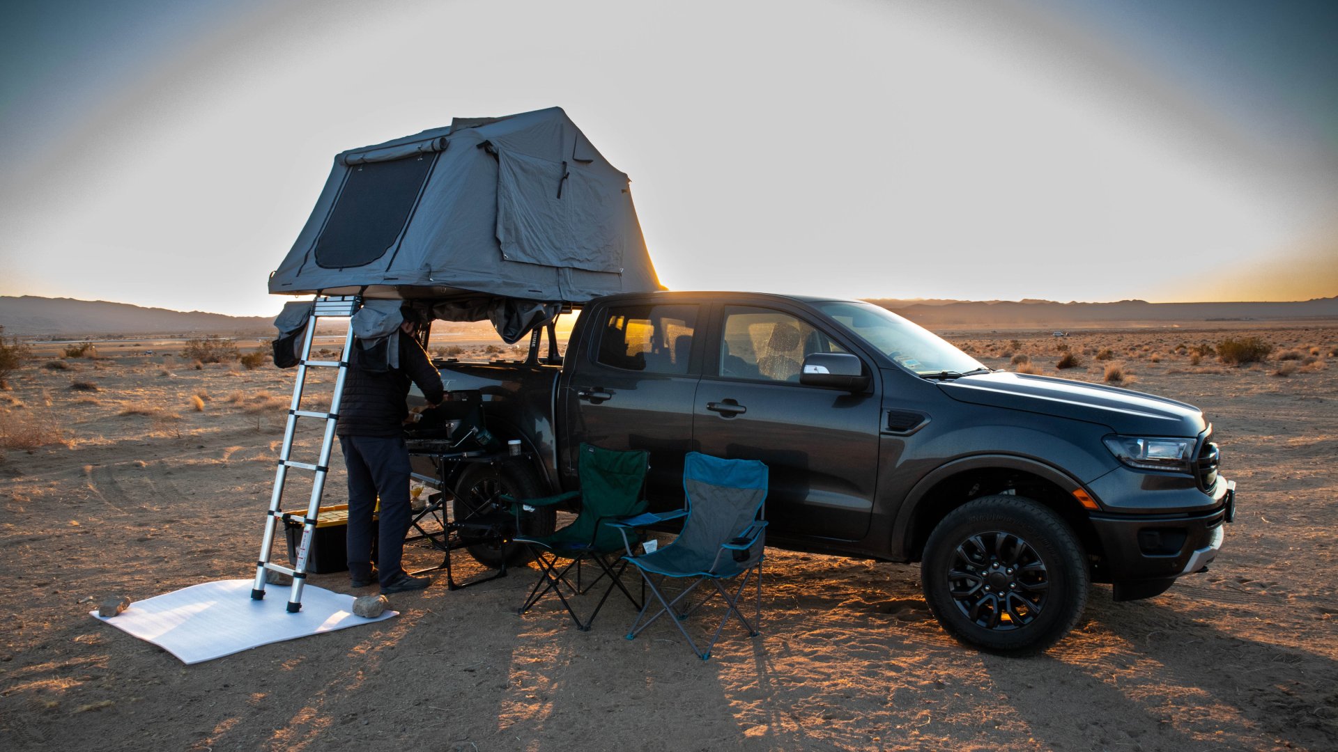 A person standing under a black pickup truck’s rooftop tent with a ladder, next to two folding chairs.